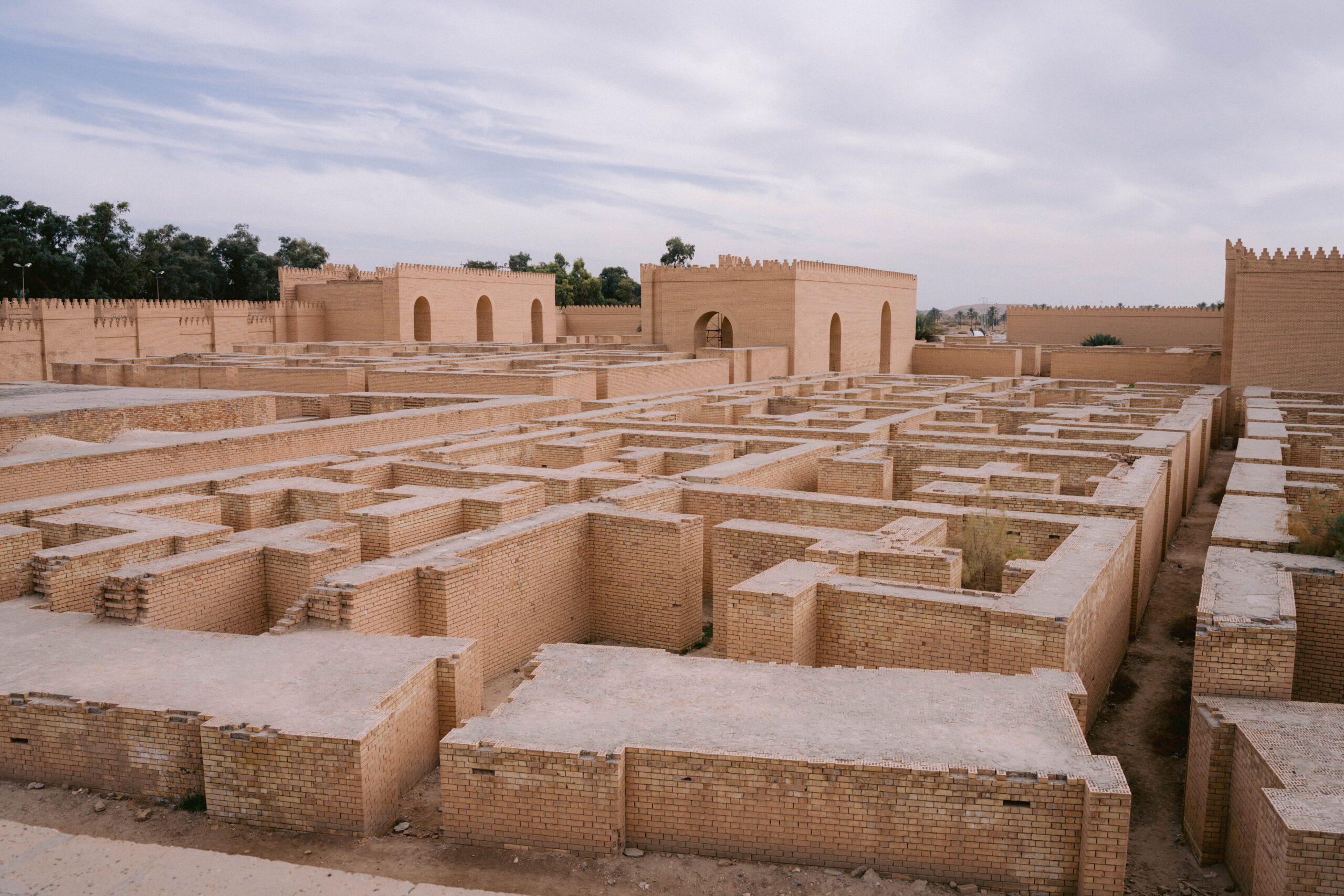 View of reconstructed ruins at the ancient city of Babylon in Iraq, showing maze-like mudbrick walls, arched gateways, and crenellated structures against a partly cloudy sky.