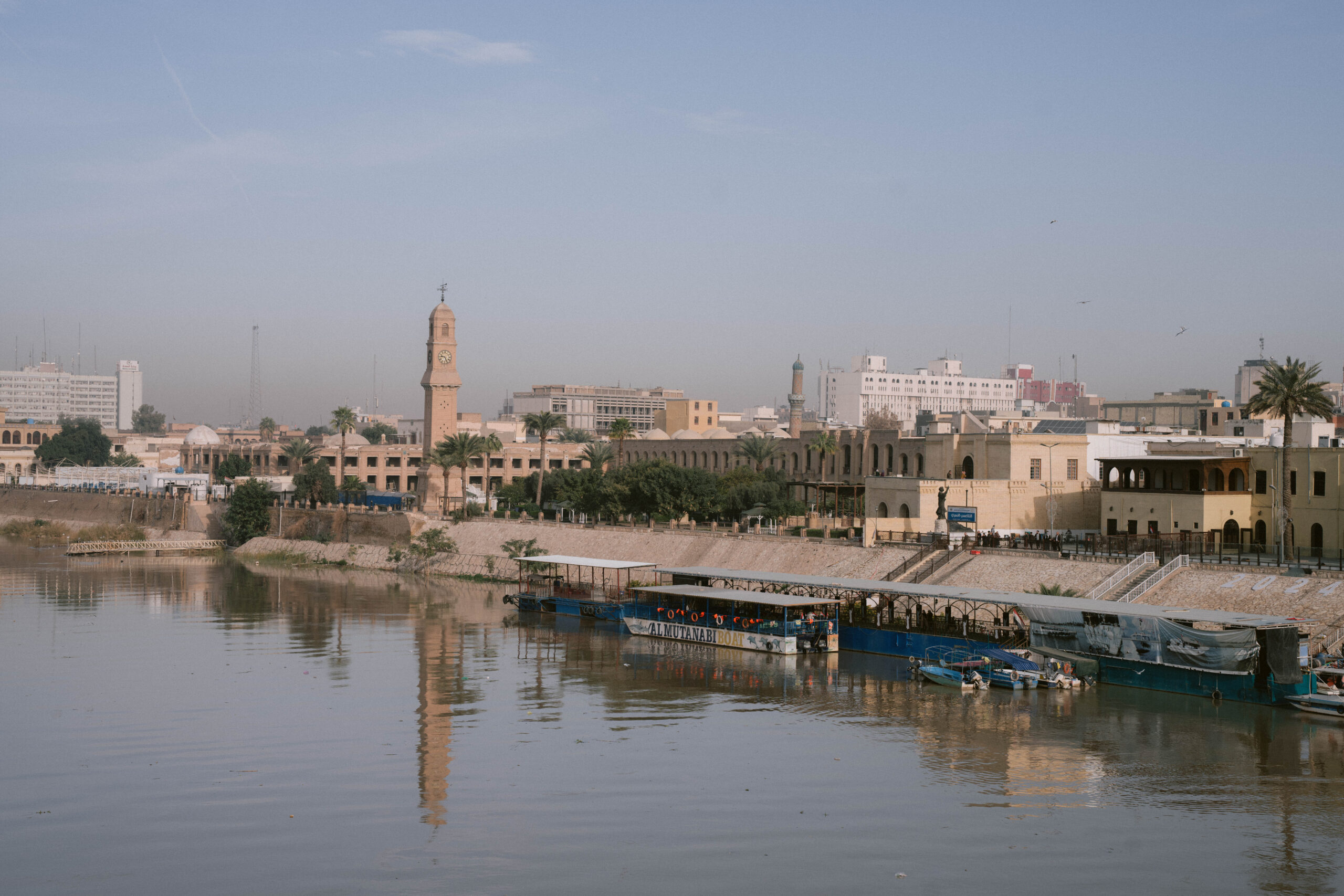 View of the Tigris River in the capital of Iraq, Baghdad, with the Al-Mutanabbi boat docked along the waterfront and the historic Ottoman-era Qishla Clock Tower rising in the background, surrounded by palm trees and traditional Iraqi architecture.