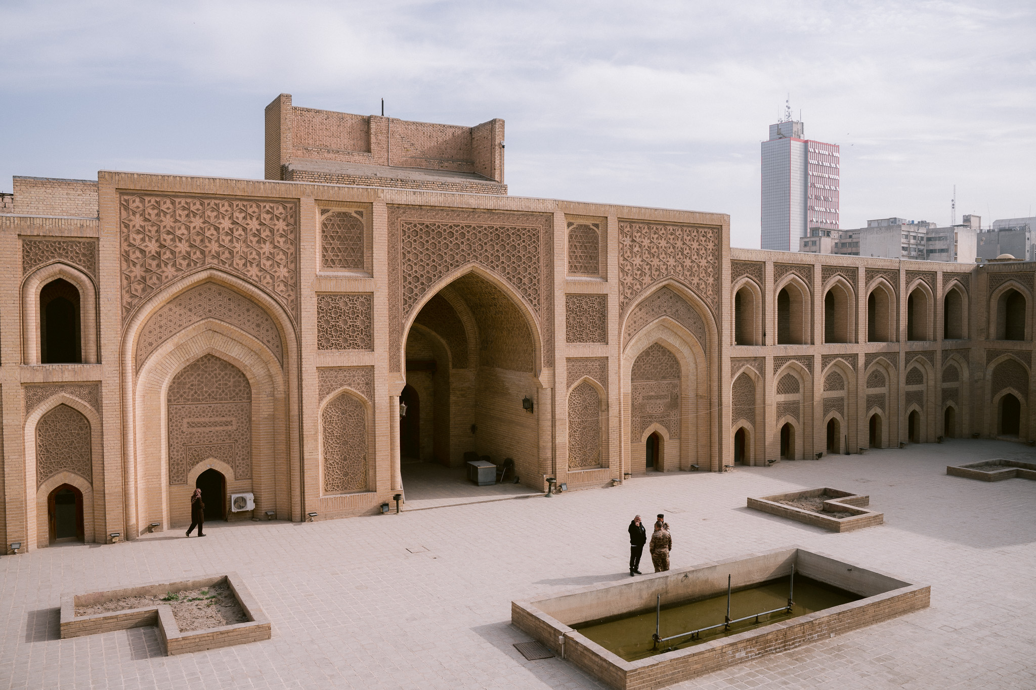Interior courtyard of the historic Al-Mustansiriya School in Baghdad, Iraq, showcasing intricate Islamic brickwork, arched doorways, and a central fountain, with a few visitors walking through the space and modern city buildings visible in the background.