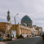 Street view of the Al-Khilani Mosque in Baghdad, Iraq, with its striking blue-tiled dome and minaret rising above surrounding buildings, as pedestrians and street vendors line the busy urban road.