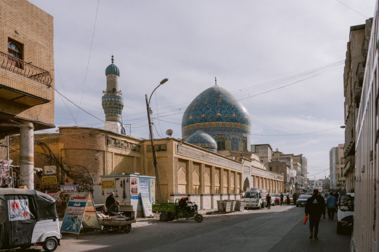 Street view of the Al-Khilani Mosque in Baghdad, Iraq, with its striking blue-tiled dome and minaret rising above surrounding buildings, as pedestrians and street vendors line the busy urban road.