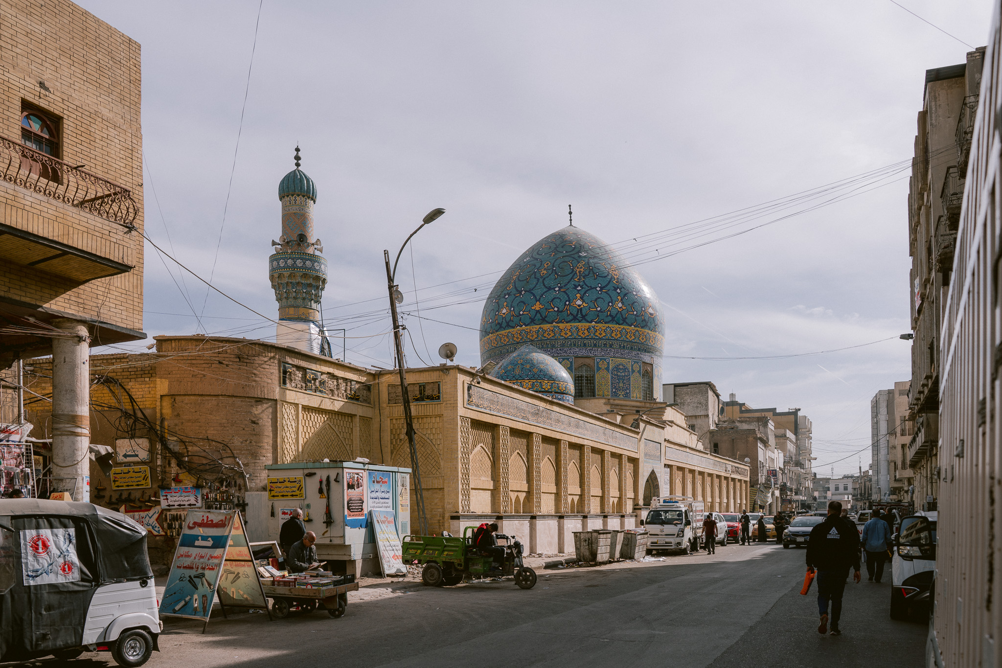 Street view of the Al-Khilani Mosque in Baghdad, Iraq, with its striking blue-tiled dome and minaret rising above surrounding buildings, as pedestrians and street vendors line the busy urban road.