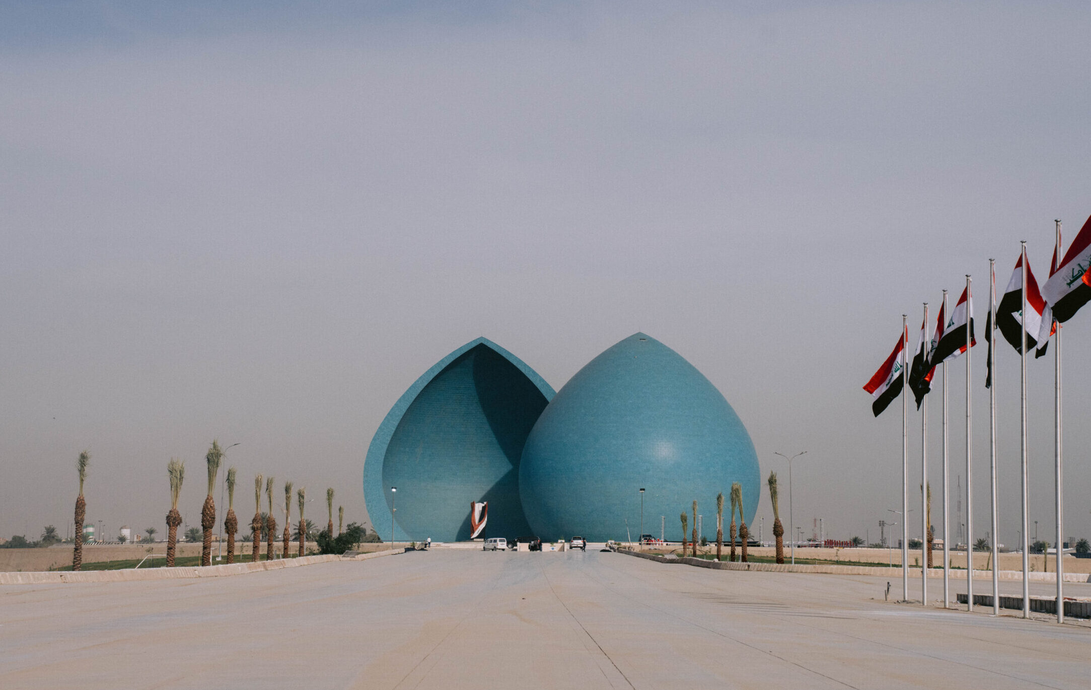 View of the Martyr’s Monument (Al-Shaheed Monument) in Baghdad, Iraq — two large blue domes split in the middle, surrounded by palm trees and Iraqi flags lining the walkway, under a hazy sky.