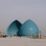 View of the Martyr’s Monument (Al-Shaheed Monument) in Baghdad, Iraq — two large blue domes split in the middle, surrounded by palm trees and Iraqi flags lining the walkway, under a hazy sky.
