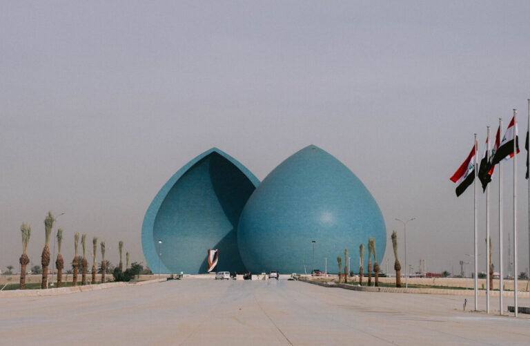 View of the Martyr’s Monument (Al-Shaheed Monument) in Baghdad, Iraq — two large blue domes split in the middle, surrounded by palm trees and Iraqi flags lining the walkway, under a hazy sky.