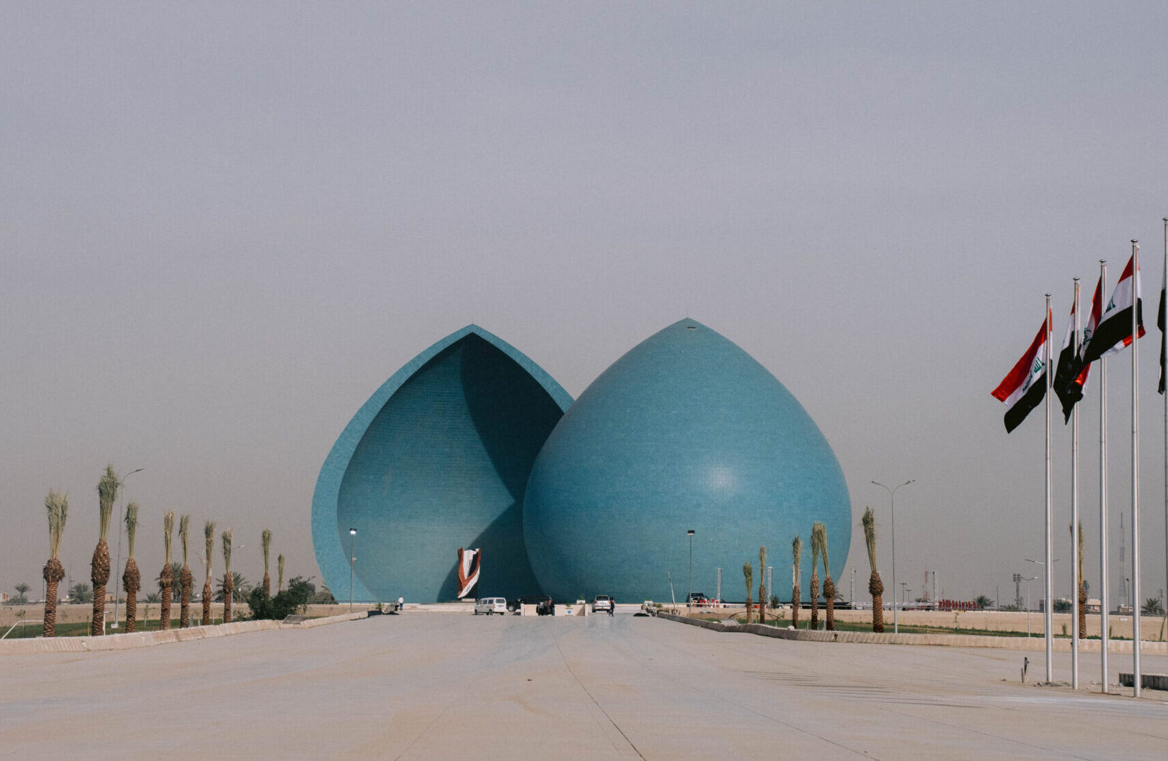 View of the Martyr’s Monument (Al-Shaheed Monument) in Baghdad, Iraq — two large blue domes split in the middle, surrounded by palm trees and Iraqi flags lining the walkway, under a hazy sky.