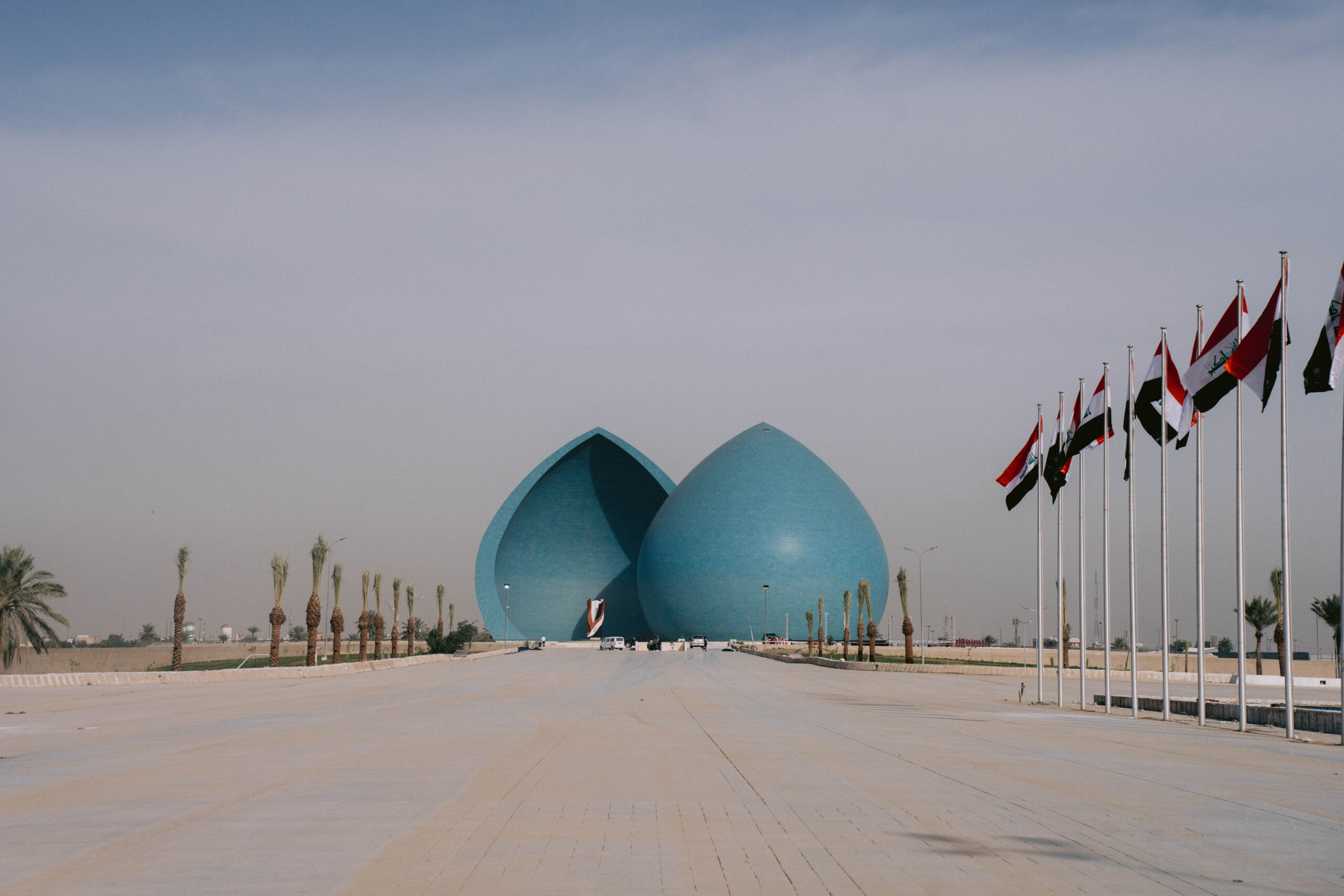 View of the Martyr’s Monument (Al-Shaheed Monument) in Baghdad, Iraq — two large blue domes split in the middle, surrounded by palm trees and Iraqi flags lining the walkway, under a hazy sky.