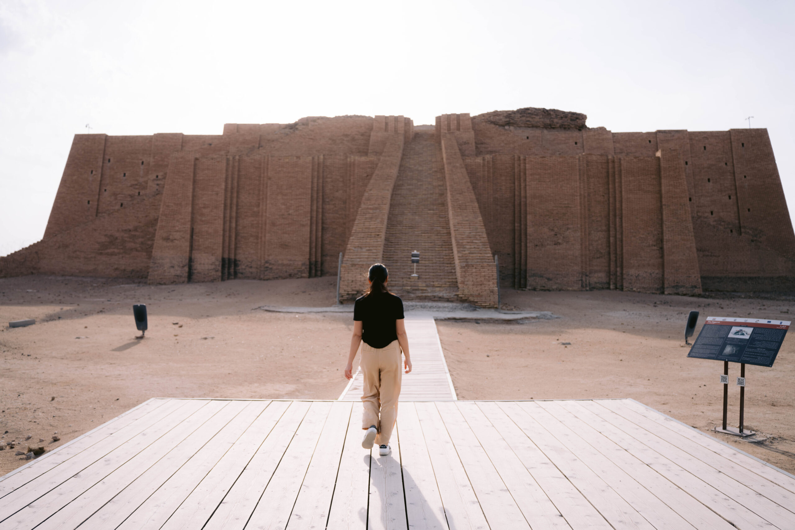 A woman walks toward the ancient Ziggurat of Ur in southern Iraq, a massive stepped structure made of mudbrick, under a bright sky. The historic site, dating back over 4,000 years, stands in a desert landscape with an information board nearby.