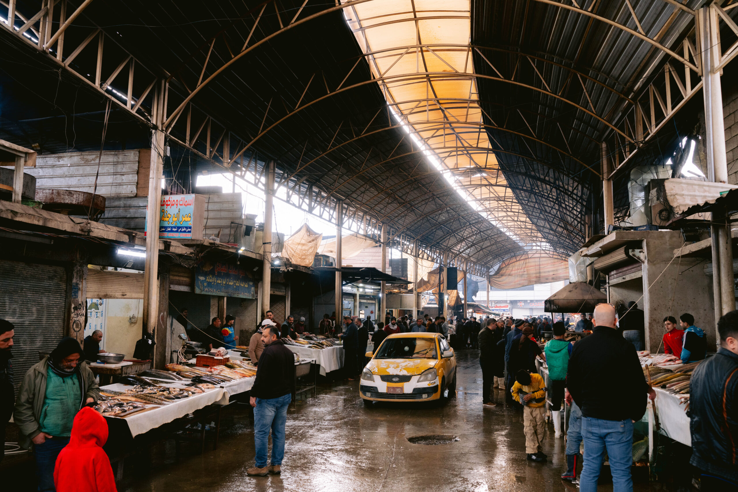 Crowded fish market in Mosul, Iraq, with vendors selling fresh seafood under a high arched roof, shoppers browsing the stalls, and a yellow taxi navigating the wet market aisle.