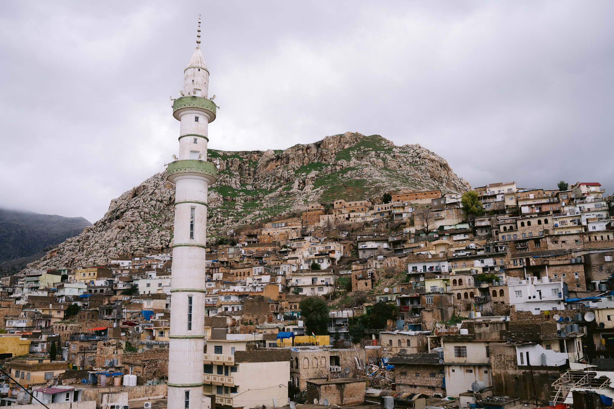 View of the mountain town of Akre in Iraqi Kurdistan, featuring a white-domed mosque with a tall minaret surrounded by hillside homes and rugged mountain peaks under a moody sky.
