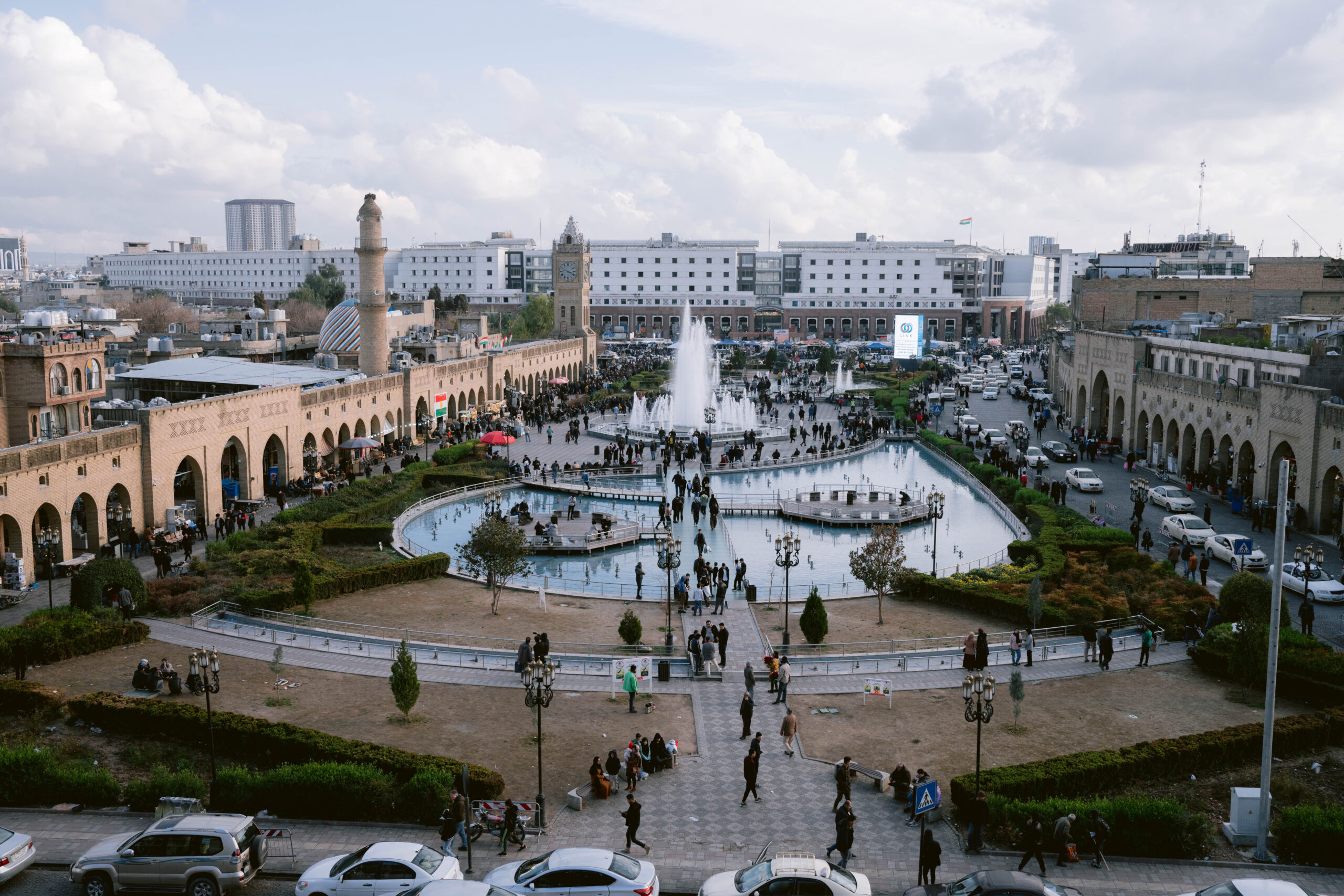 Erbil’s bustling city center in Iraqi Kurdistan, featuring the large fountains of Shanidar Park, surrounded by crowds, cars, and the historic Qaysari Bazaar arcade with its iconic clock tower in the background.