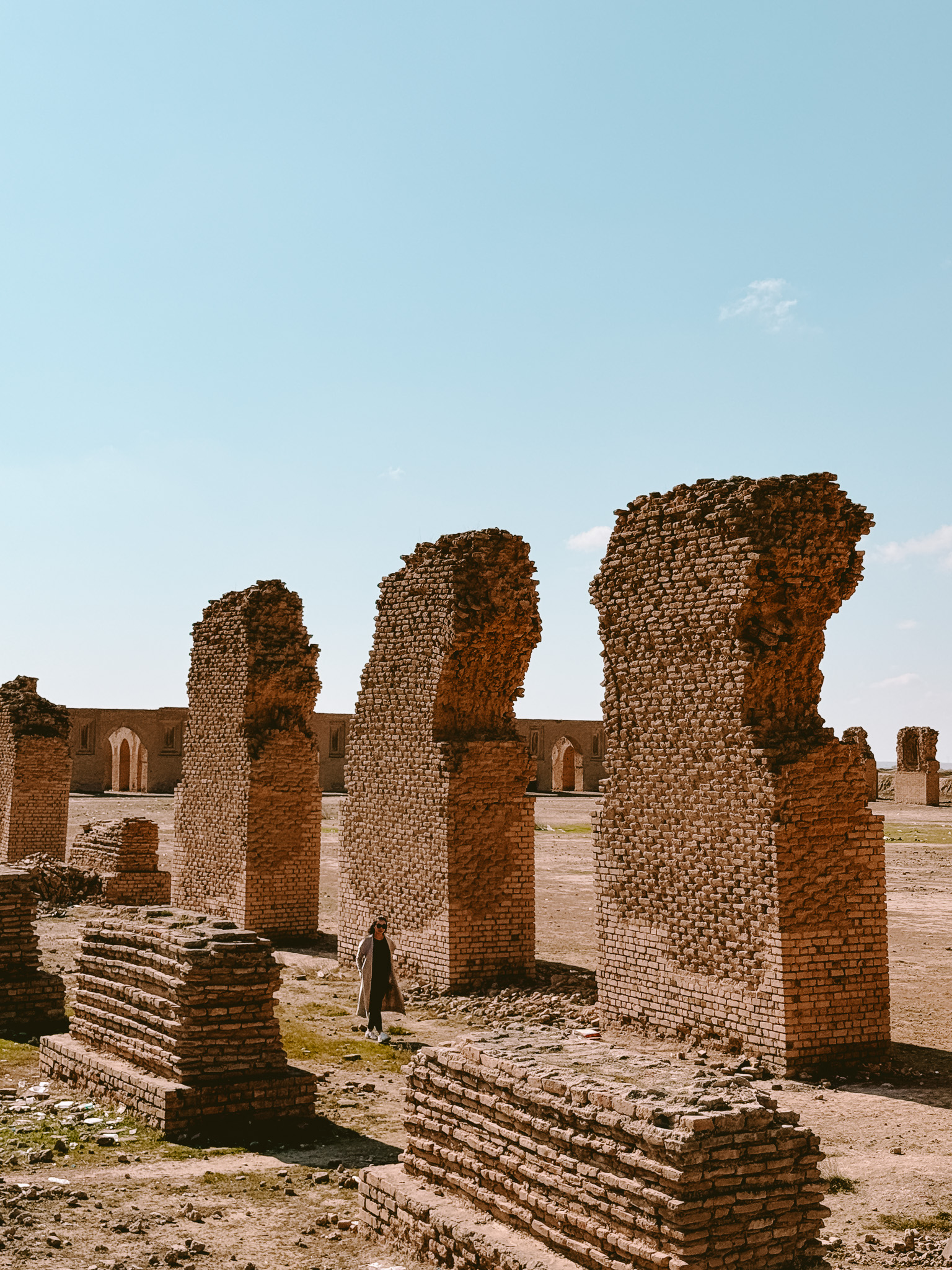 A woman walks among the crumbling mudbrick arches of the ancient Abbasid-era ruins in Samarra, Iraq, surrounded by weathered stone blocks under a clear blue sky.