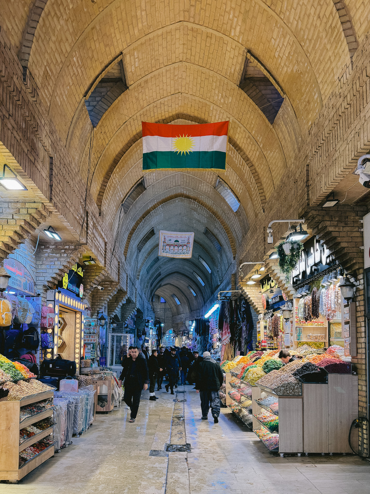 Interior view of Erbil’s Qaysari Bazaar in Iraqi Kurdistan, with shoppers walking beneath arched brick ceilings and colorful market stalls selling candy, textiles, and goods, while the Kurdish flag hangs overhead.