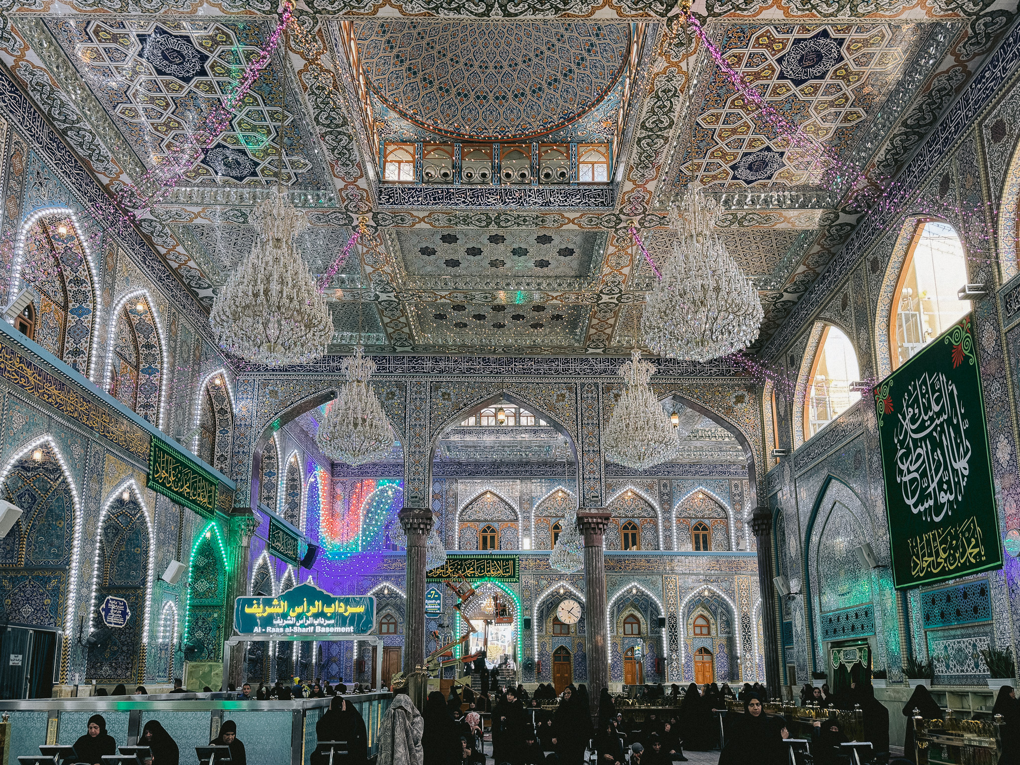 Interior of the Imam Ali Shrine in Najaf, Iraq, showcasing intricate mirrored mosaics, ornate tilework, grand chandeliers, and worshippers gathered beneath the decorated ceilings and arches in prayer.