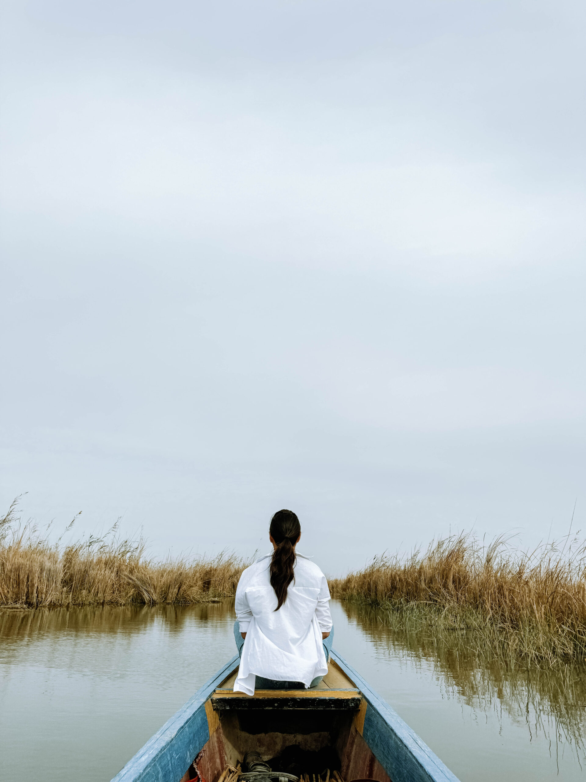 A woman sits at the front of a wooden canoe gliding through the reed-lined waters of the Mesopotamian Marshes in southern Iraq, facing forward under an overcast sky.