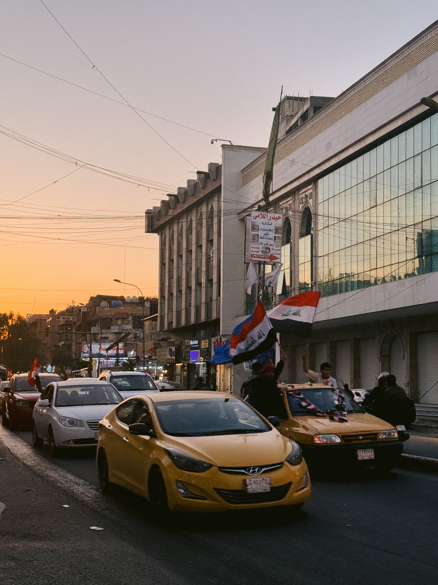 Street scene at sunset in Iraq with young men waving Iraqi flags from yellow taxis, likely during a celebration or protest. The street is lined with modern buildings, Arabic signage, and a reflection of the sky in a glass façade.