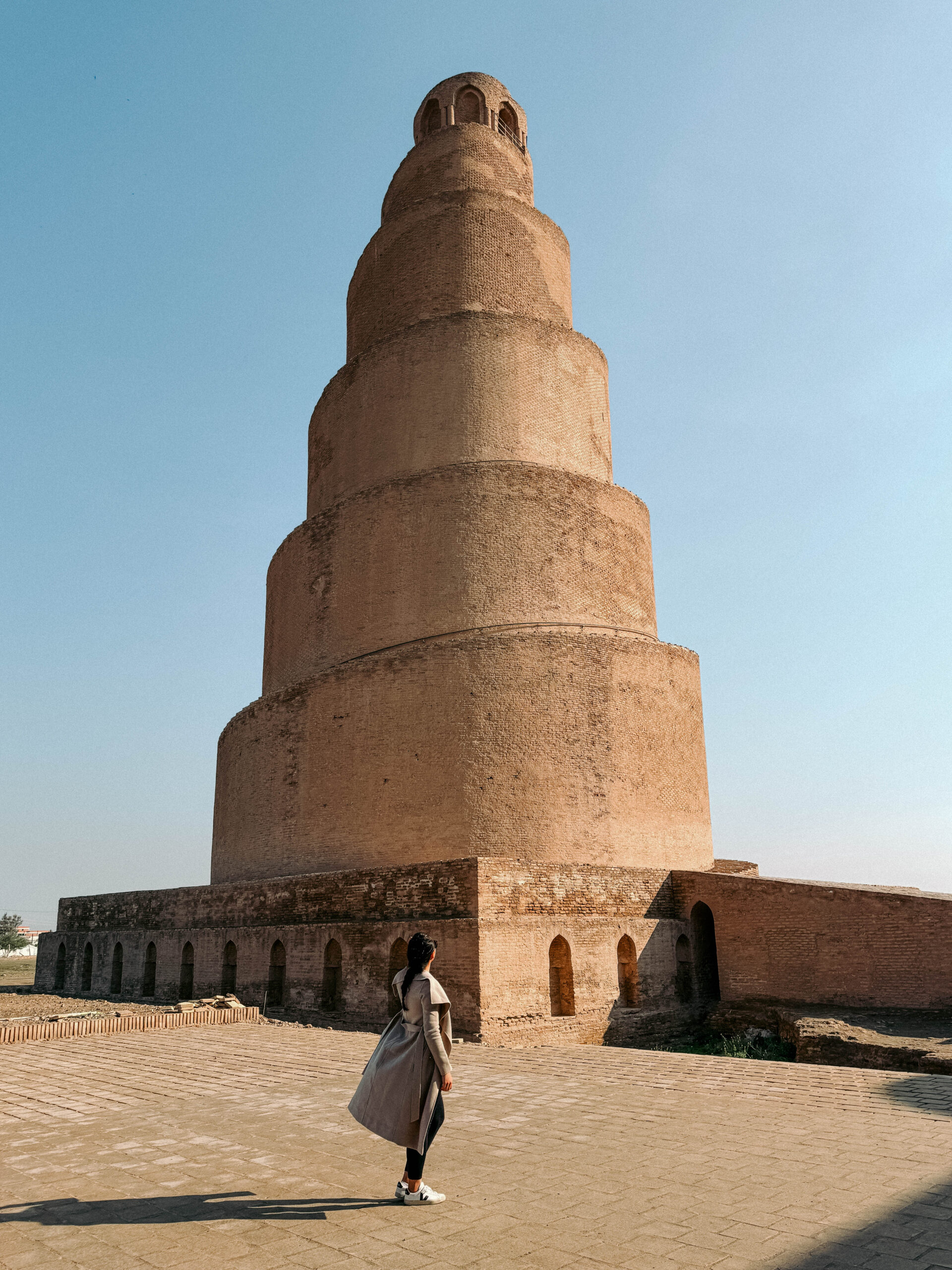 A woman stands in front of the towering spiral minaret of the Great Mosque of Samarra in Iraq, gazing up at the 9th-century Malwiya Minaret under a clear blue sky.