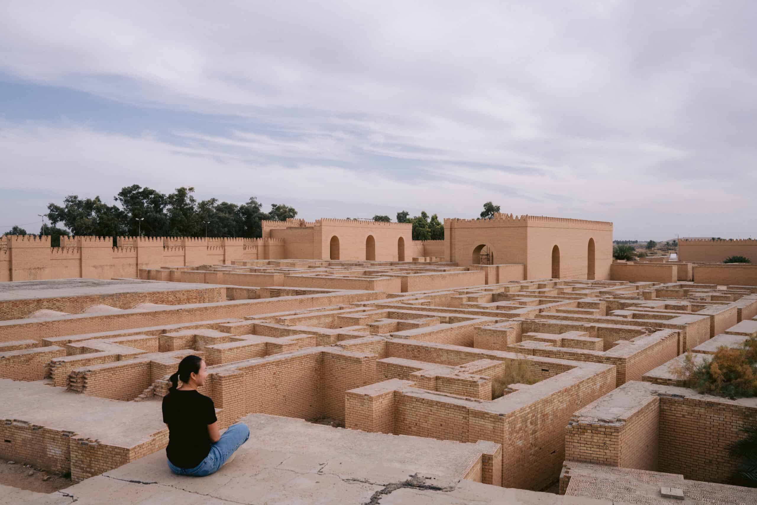 Traveler sitting on a ledge overlooking the reconstructed maze-like walls of ancient Babylon, Iraq, with fortified gateways and brick architecture in the background.