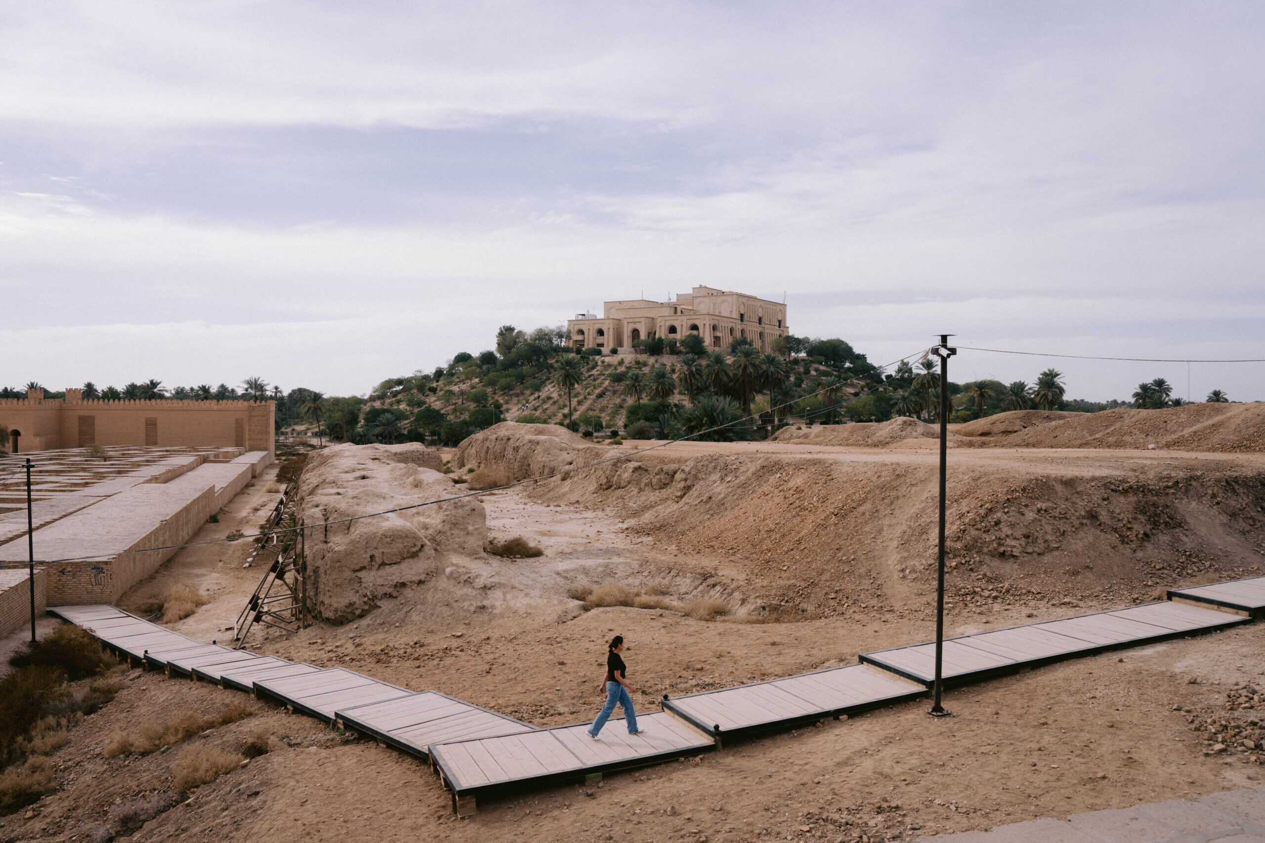 A visitor walks along a boardwalk through the ancient ruins of Babylon, Iraq, with Saddam Hussein’s former palace visible on the hilltop in the background.