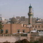 View from a bridge over the Tigris River in Baghdad, Iraq, showing a vibrant mosque dome and minaret surrounded by city buildings and birds in flight—capturing the everyday beauty of Baghdad beyond the landmarks.