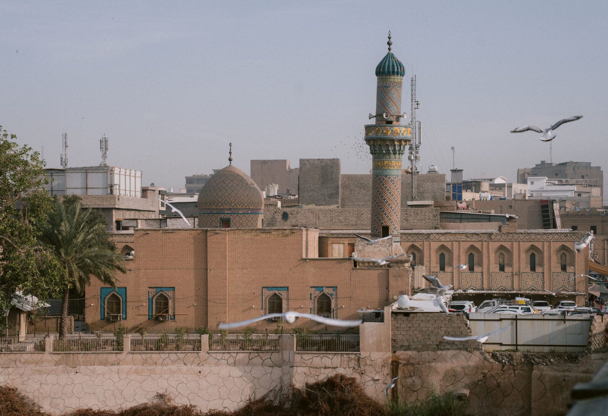 View from a bridge over the Tigris River in Baghdad, Iraq, showing a vibrant mosque dome and minaret surrounded by city buildings and birds in flight—capturing the everyday beauty of Baghdad beyond the landmarks.