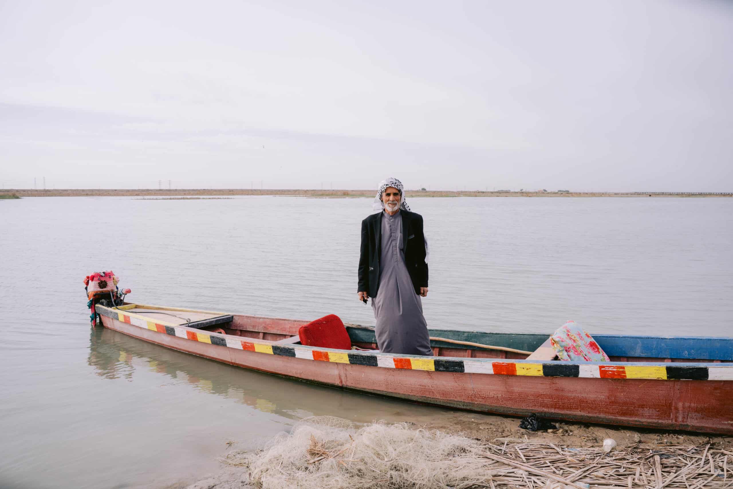 Local Marsh Arab man standing beside his colorful wooden canoe on the edge of the Mesopotamian Marshes in southern Iraq, with fishing nets in the foreground.