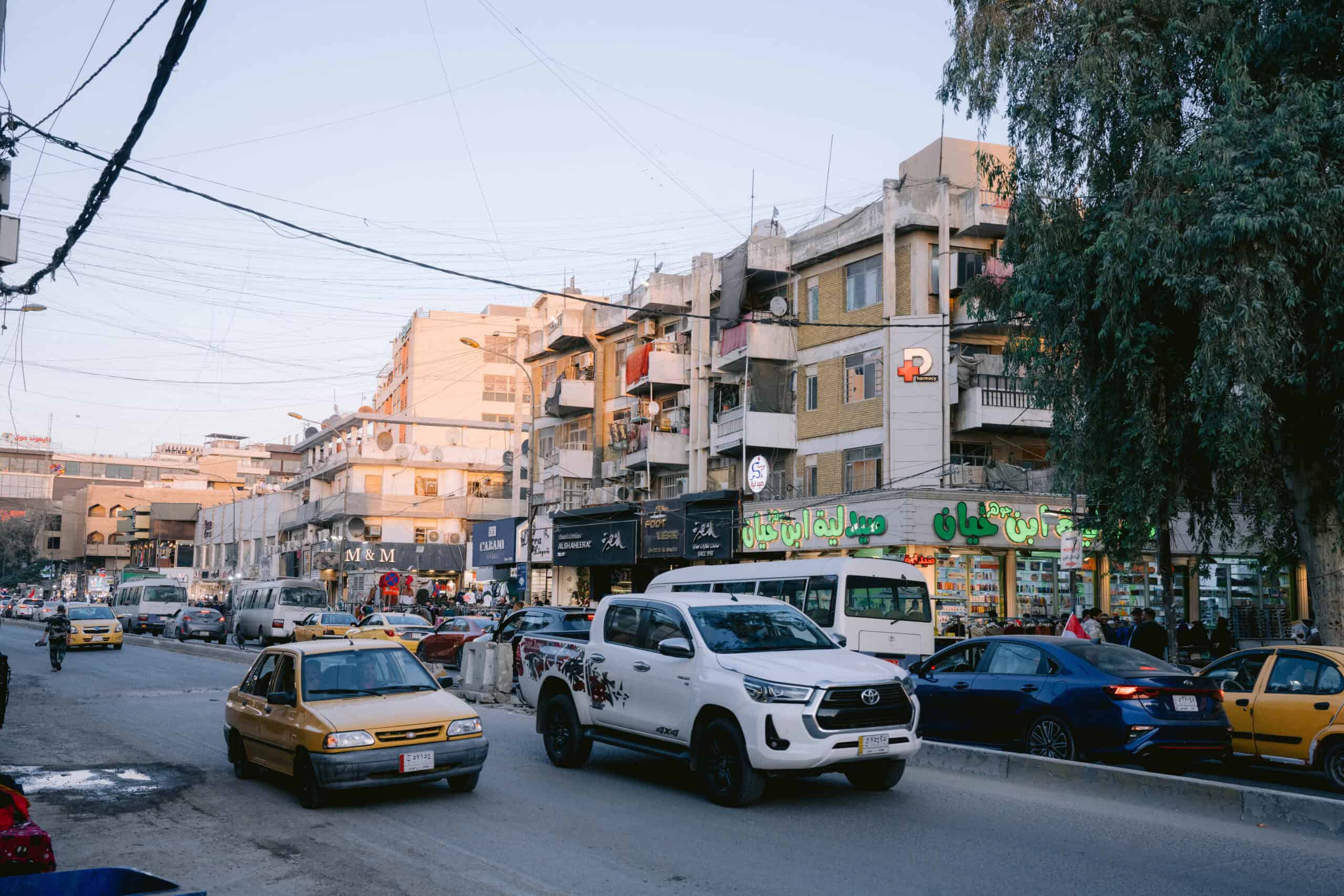 Street scene in Baghdad, Iraq at sunset, showing busy traffic with yellow taxis, storefronts with Arabic signage, pedestrians, and weathered apartment buildings under a tangle of overhead wires.