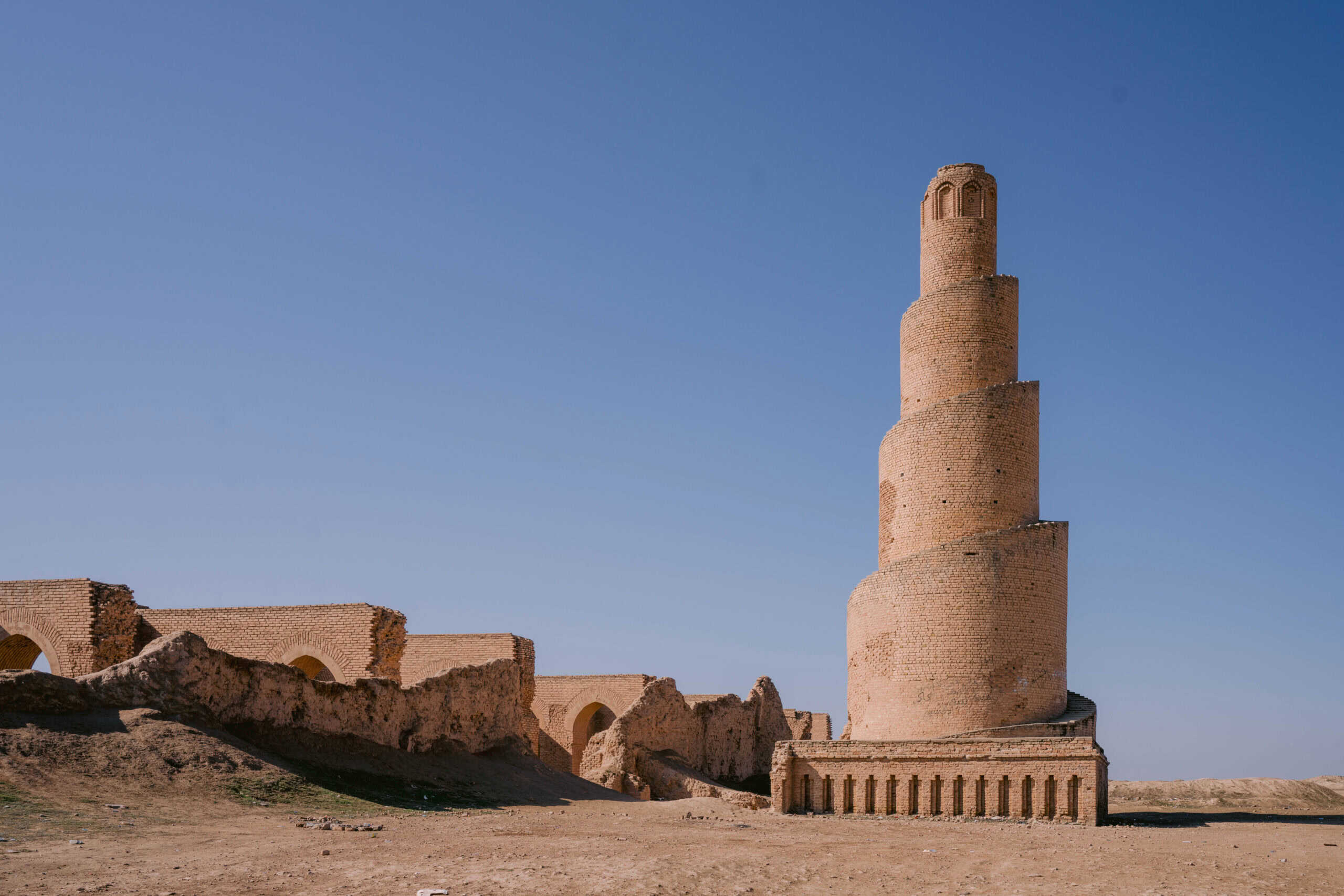 The spiral minaret of the Abu Dulaf Mosque in Iraq, made of sand-colored bricks rising against a clear blue sky, with ruins of the mosque complex surrounding its base.