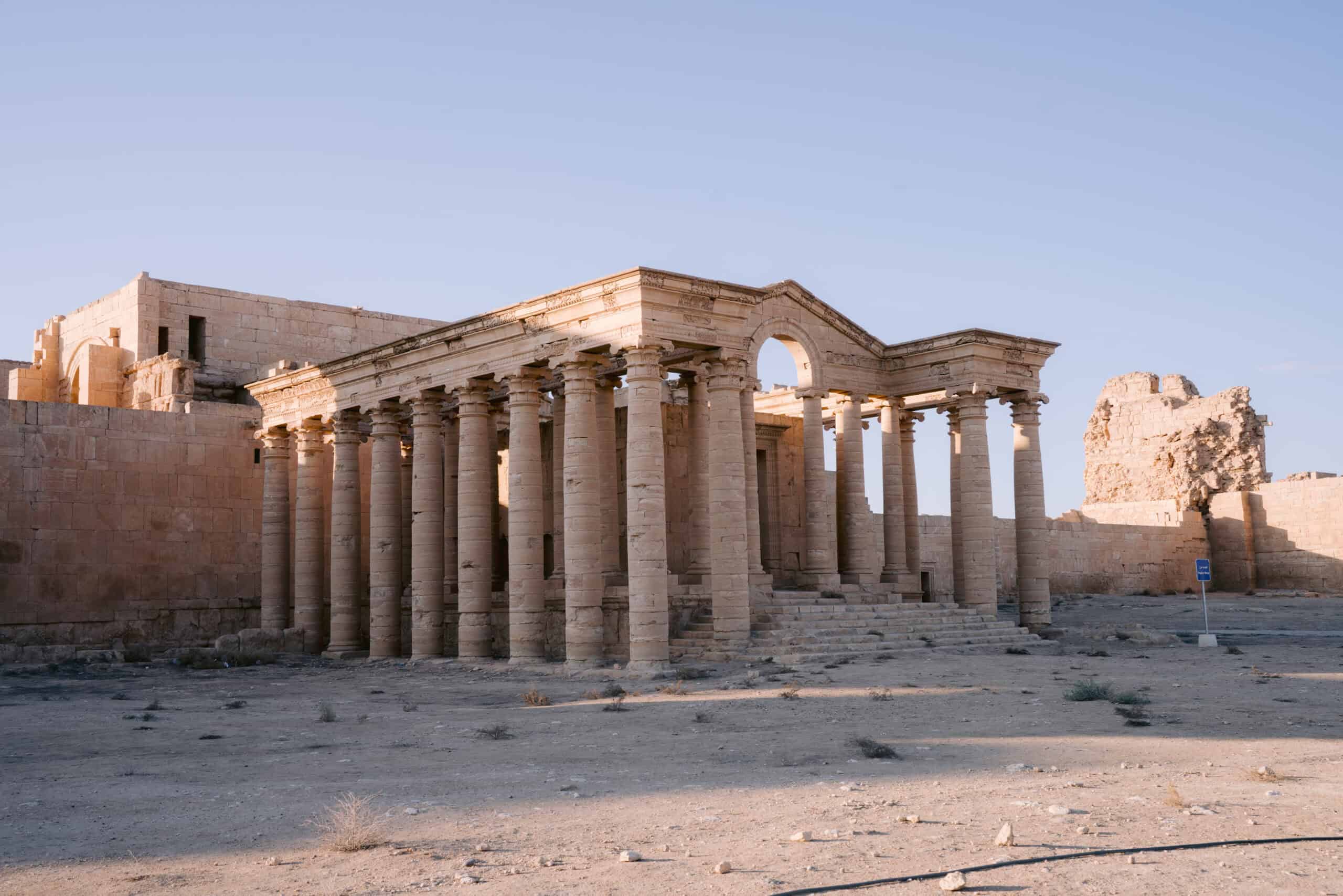 A sunlit Greco-Roman colonnaded structure with weathered stone columns and ruins in a remote desert archaeological site.