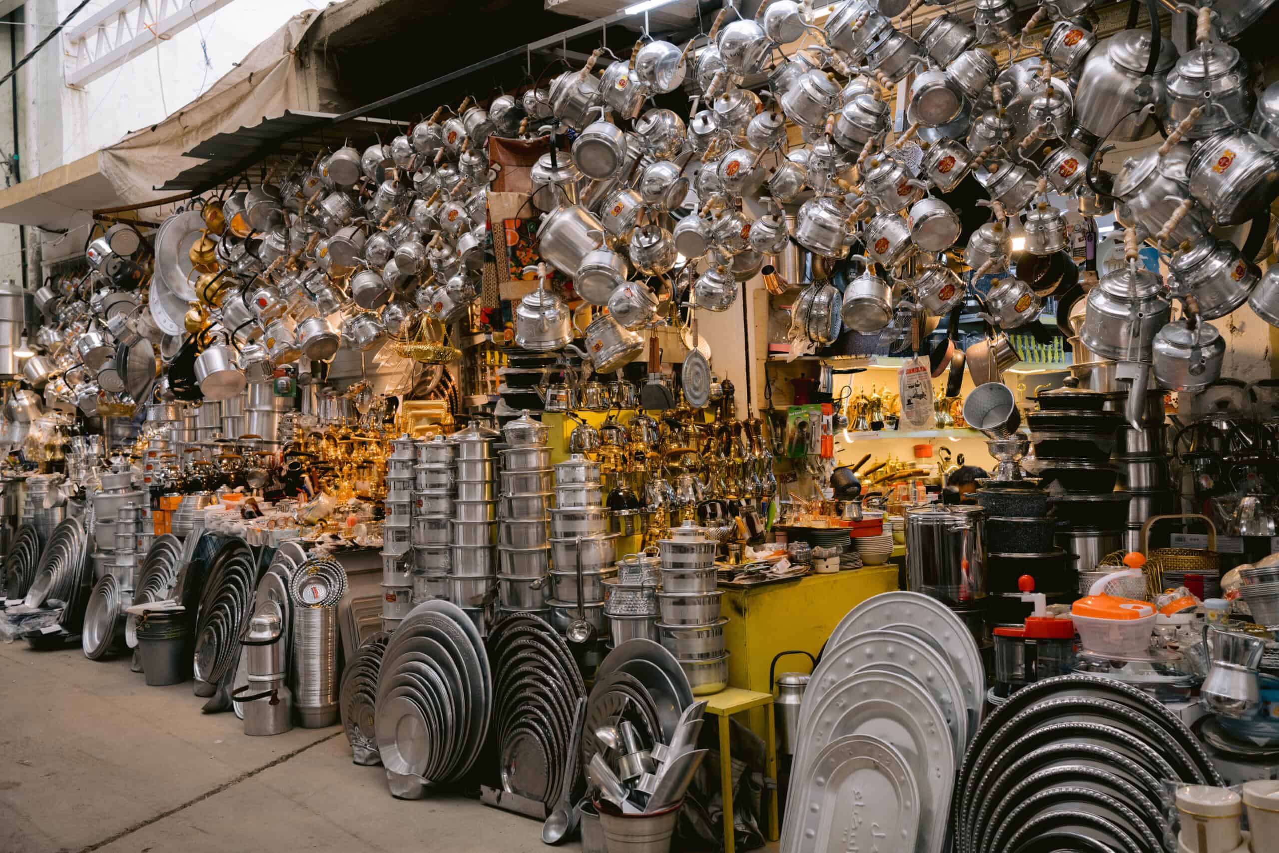 Market stall in Iraq overflowing with shiny metal kitchenware—kettles, pots, pans, and trays—stacked and hanging in organized chaos, creating a dazzling wall of silver under fluorescent lighting.