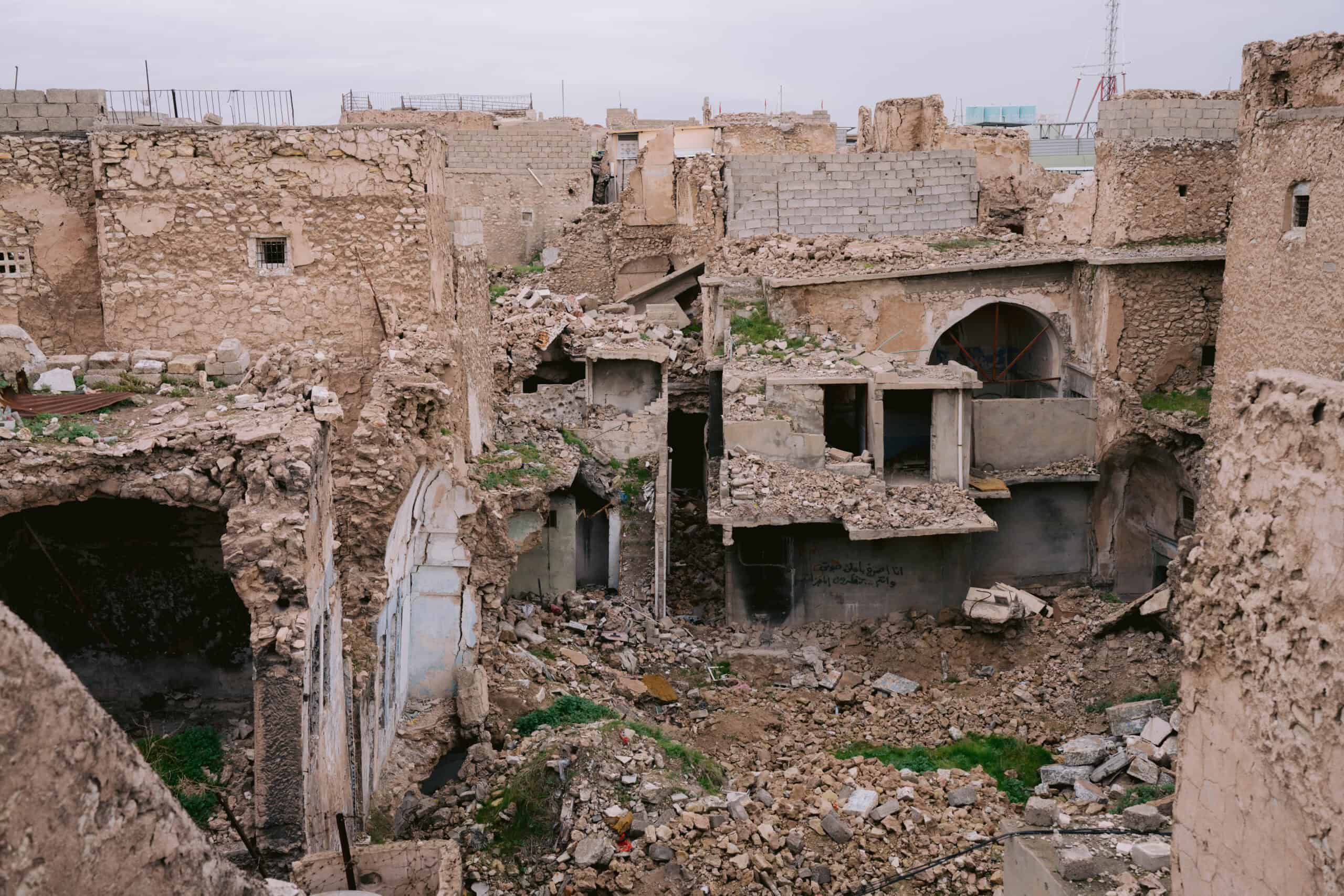 War-torn buildings in Mosul’s old city, with collapsed stone walls, piles of rubble, and remnants of homes showing visible damage from past conflict.