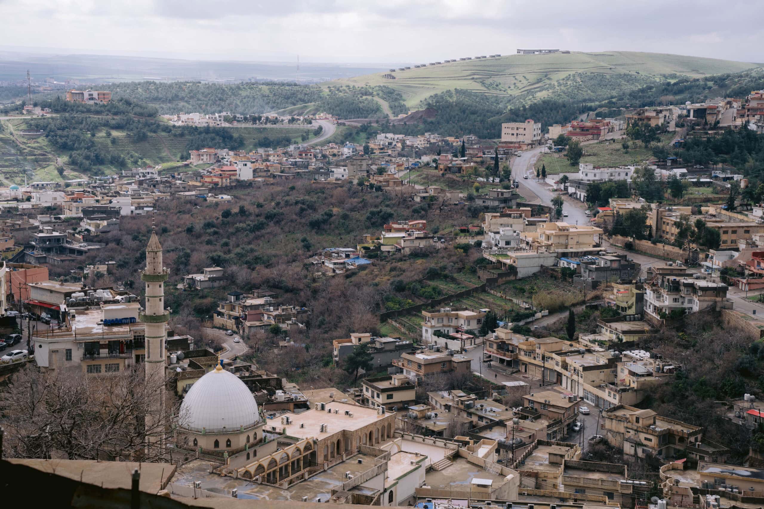 Panoramic view of Akre, Iraq, with terraced homes climbing the hills, a white-domed mosque in the foreground, and green mountains stretching into the distance under a cloudy sky.
