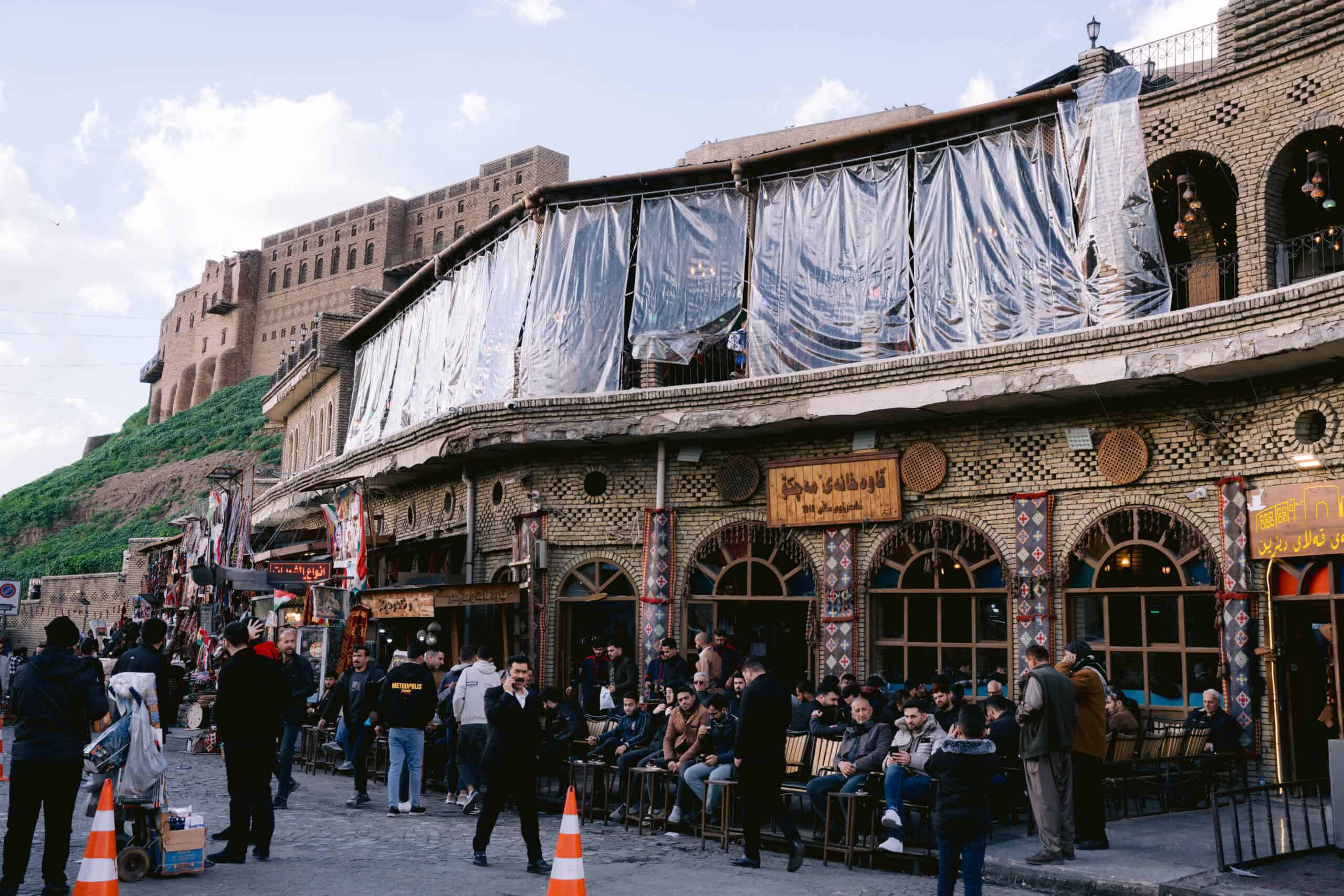 Crowded tea house with traditional brick architecture and colorful tiles in front of Erbil Citadel, with people gathered on the sidewalk and plastic coverings draped over the second-floor windows.