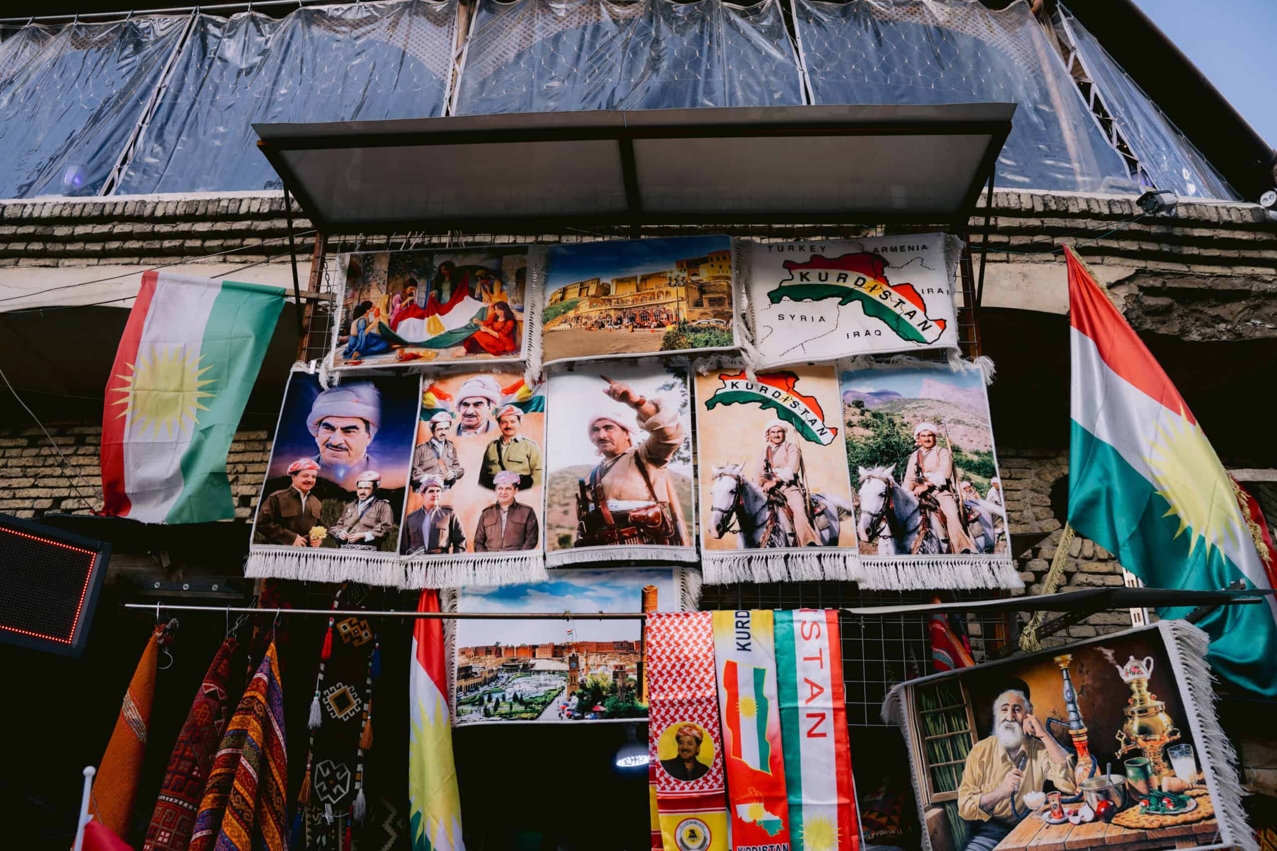 A stall displays vibrant Kurdish flags and tapestries featuring historical Kurdish leaders, traditional scenes, and political maps.