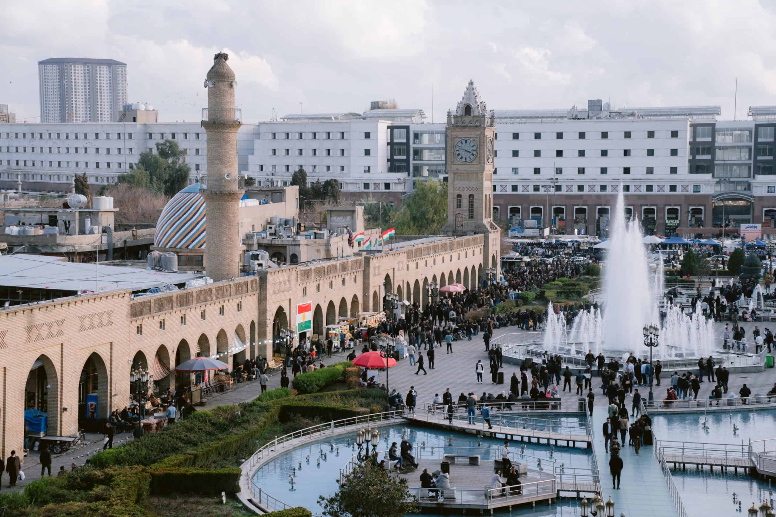 Fountains shoot into the air in a crowded square near the Erbil Citadel, surrounded by arched colonnades, a clock tower, and modern buildings.
