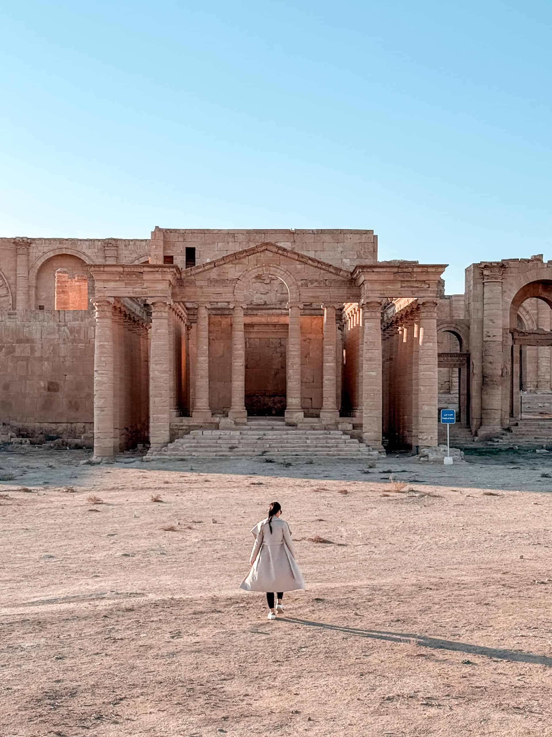 A woman in a long coat walks across a dusty clearing toward the monumental columns and facade of the ruins at Hatra.
