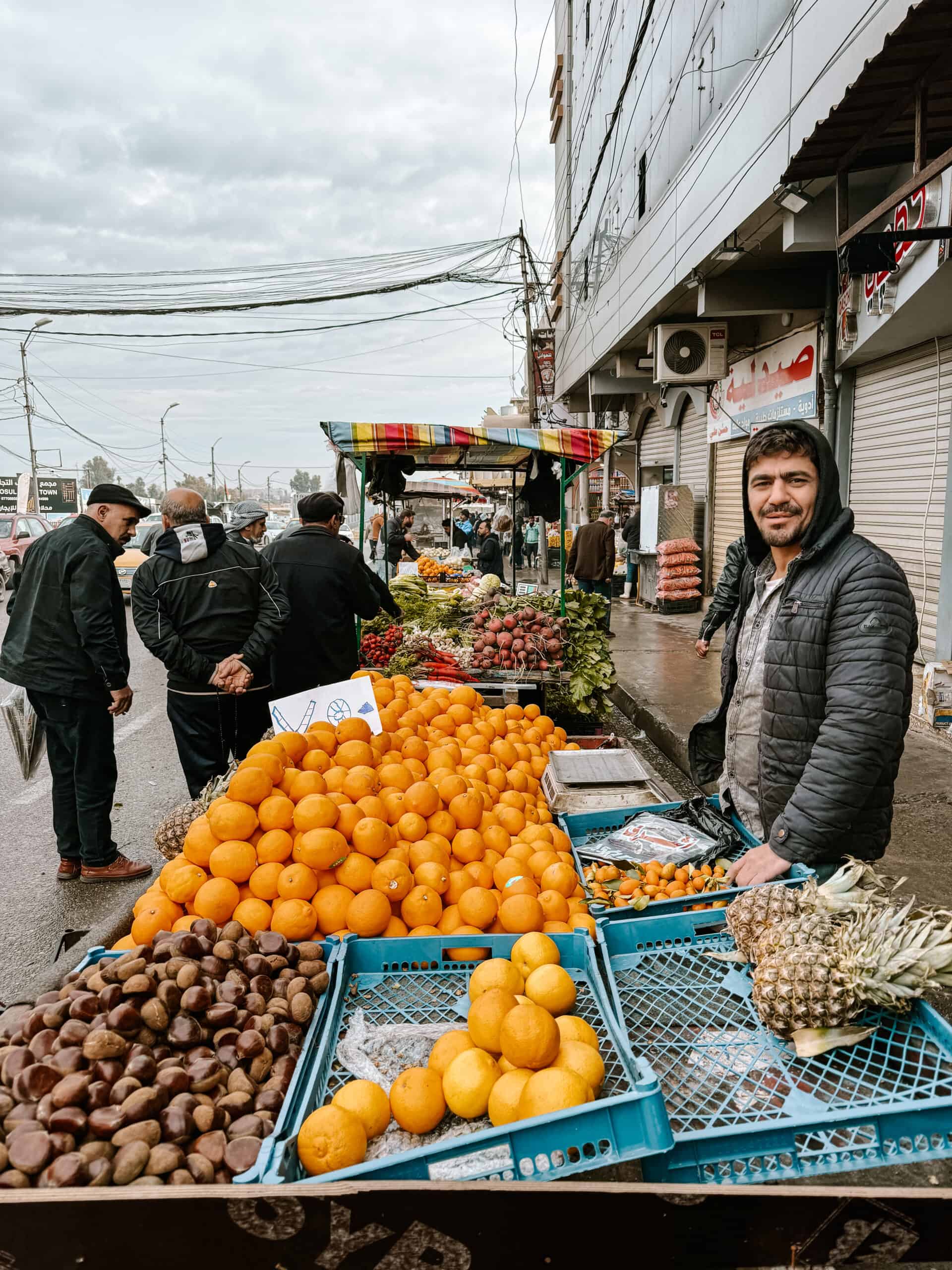A fruit vendor smiles at the camera beside stacks of oranges, pineapples, and chestnuts at a street market with passing pedestrians and nearby shops.
