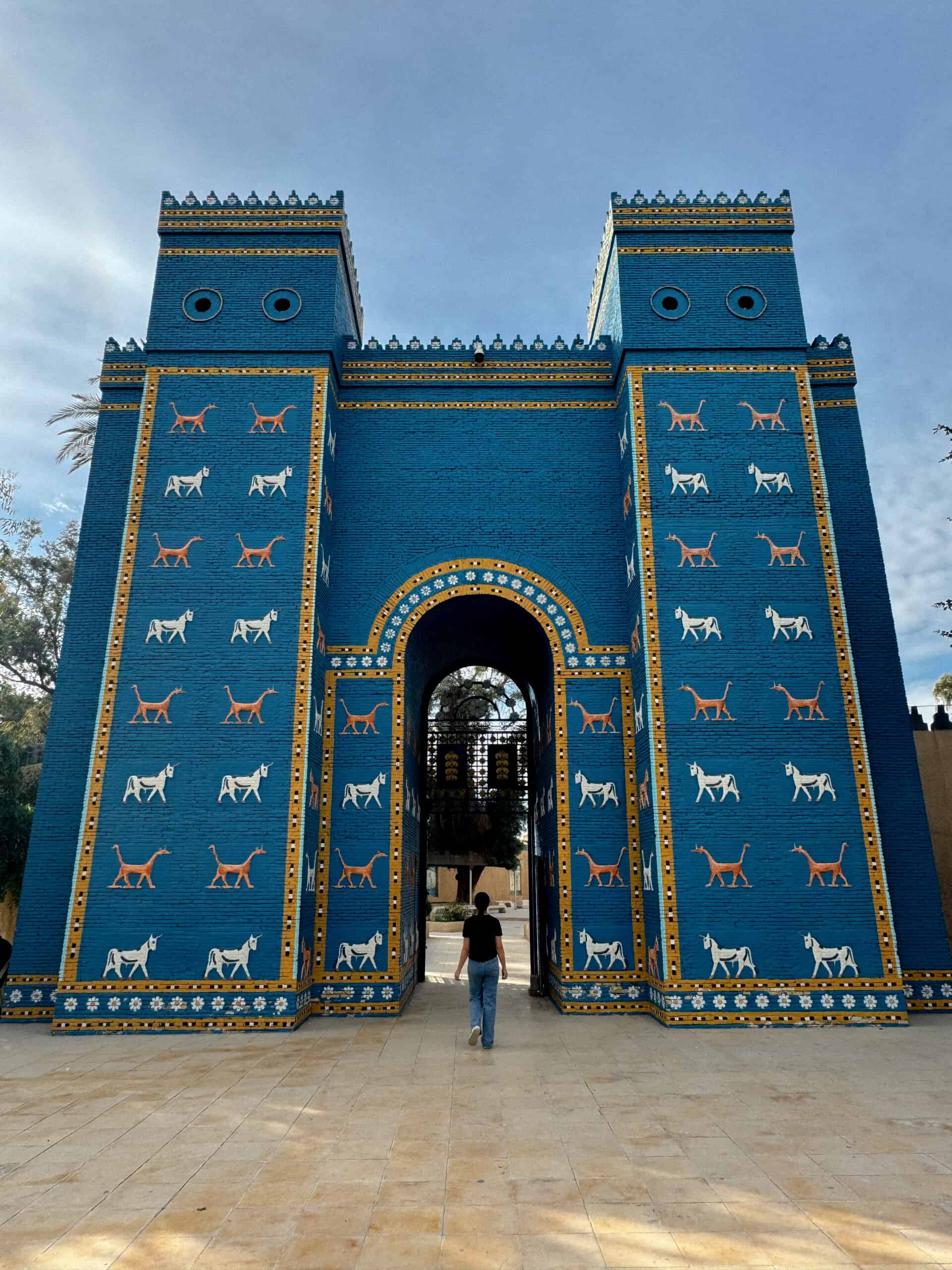 A person walks toward a reconstructed Ishtar Gate, painted bright blue and adorned with rows of yellow and white animal reliefs from ancient Babylon.