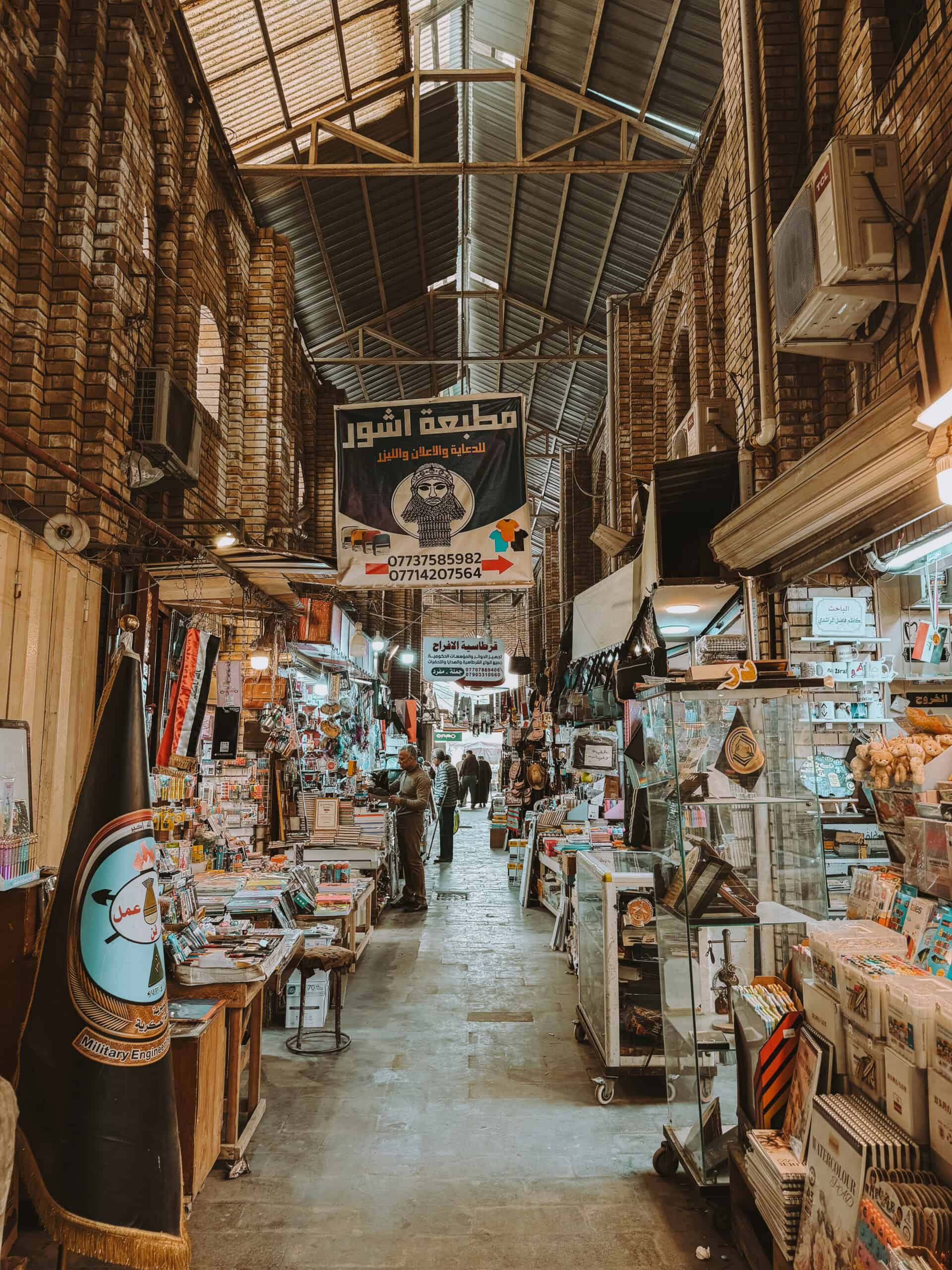 A covered bazaar corridor lined with tightly packed bookstore stalls, stationery items, and hanging banners under a metal roof.