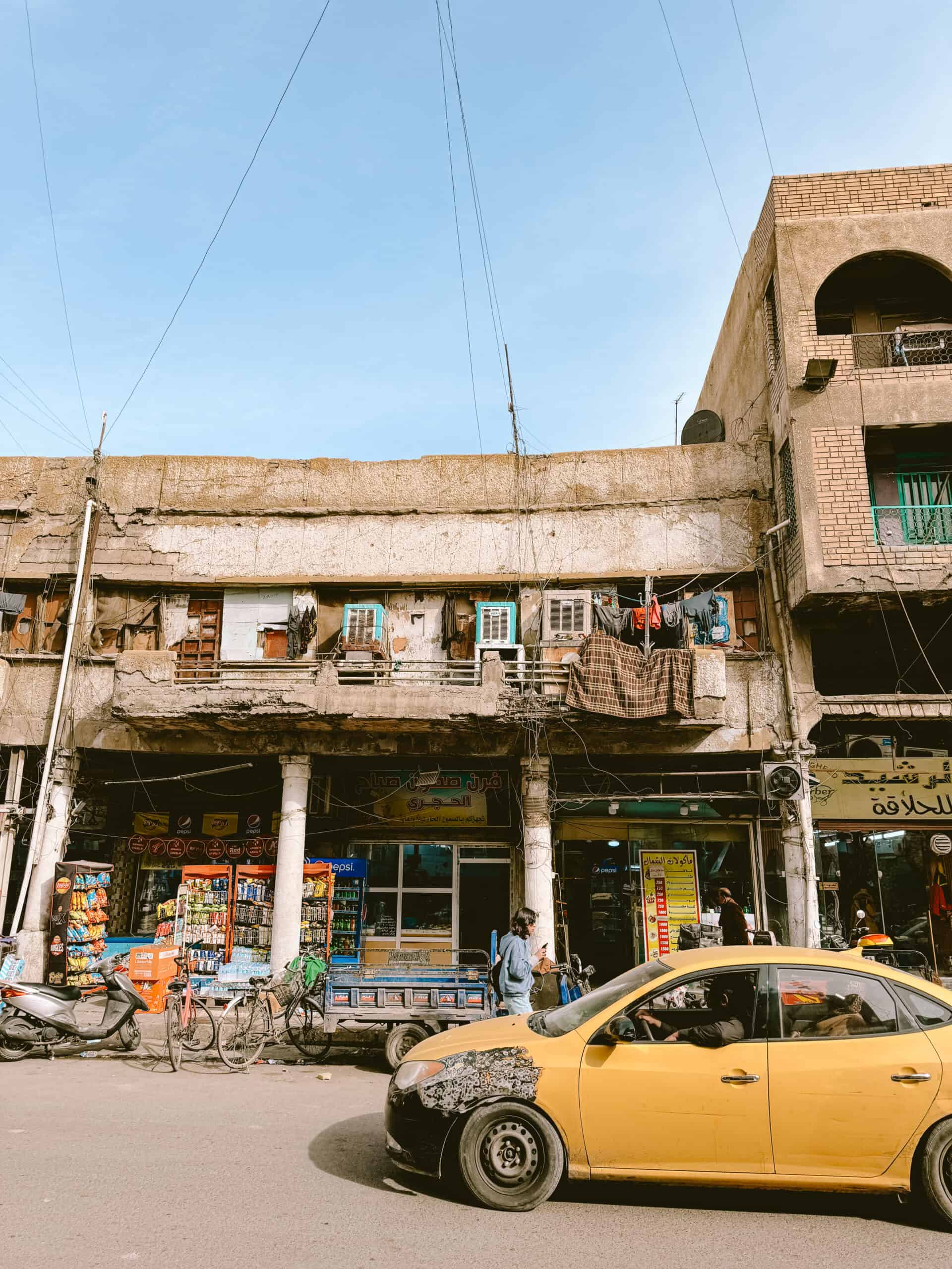 Street scene in Iraq showing a weathered two-story building with small shops at street level, snacks and drinks displayed outside, laundry hanging from the upper floor, and a worn yellow taxi driving past.