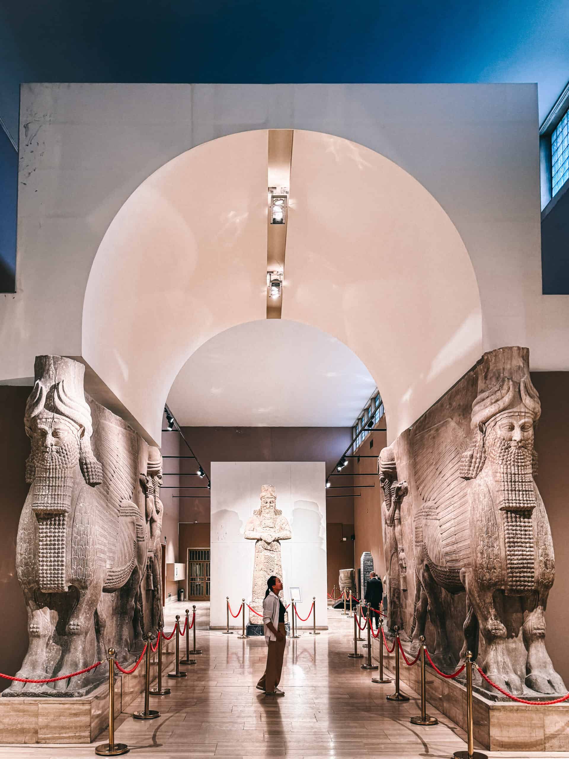 A visitor stands beneath a large stone arch flanked by two monumental Lamassu statues at the Iraq Museum in Baghdad, with an ancient Assyrian relief visible in the background.