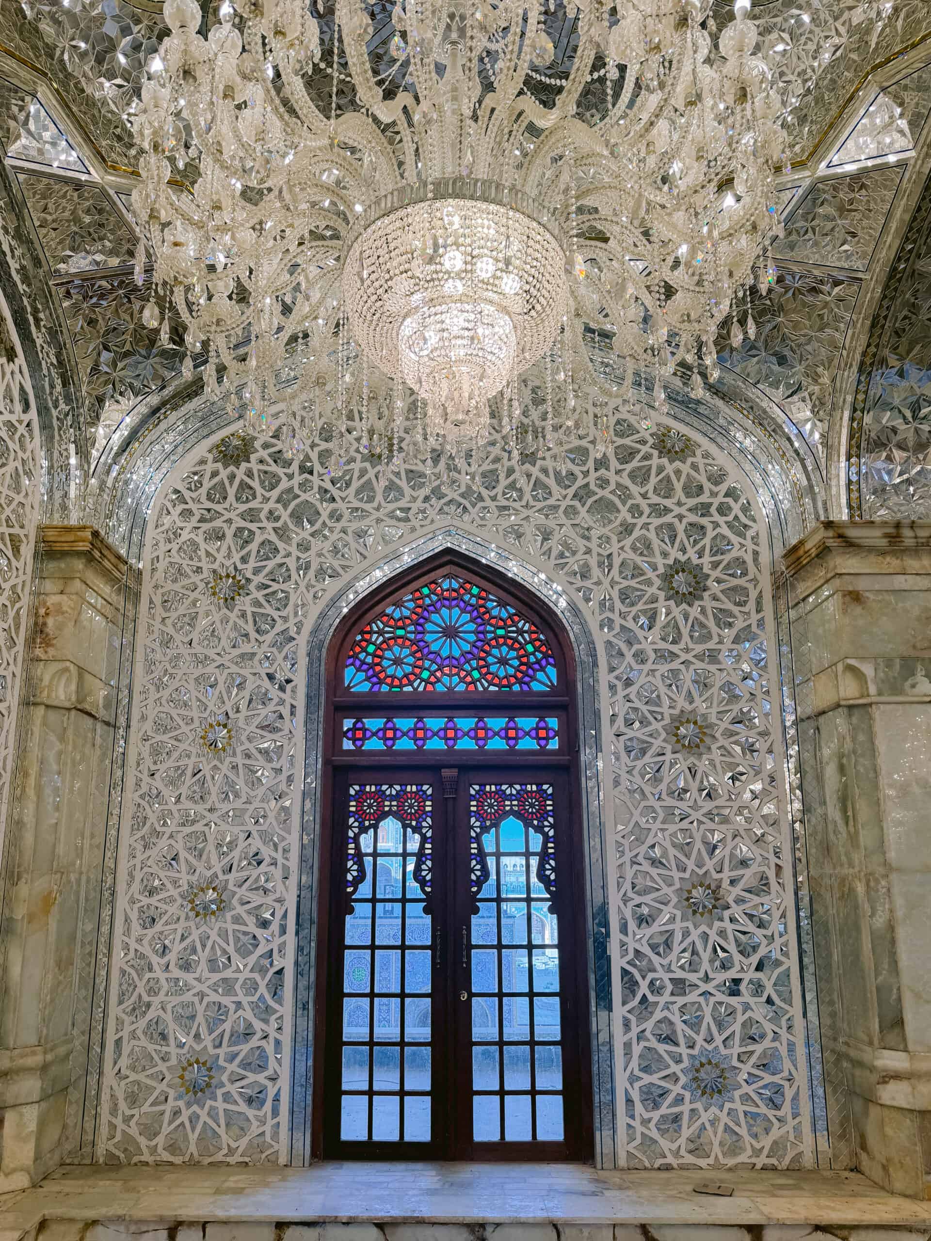 Interior of a shrine adorned with mirrored mosaic tiles, a crystal chandelier, and a stained glass window with geometric Islamic patterns and ornate wooden framing.
