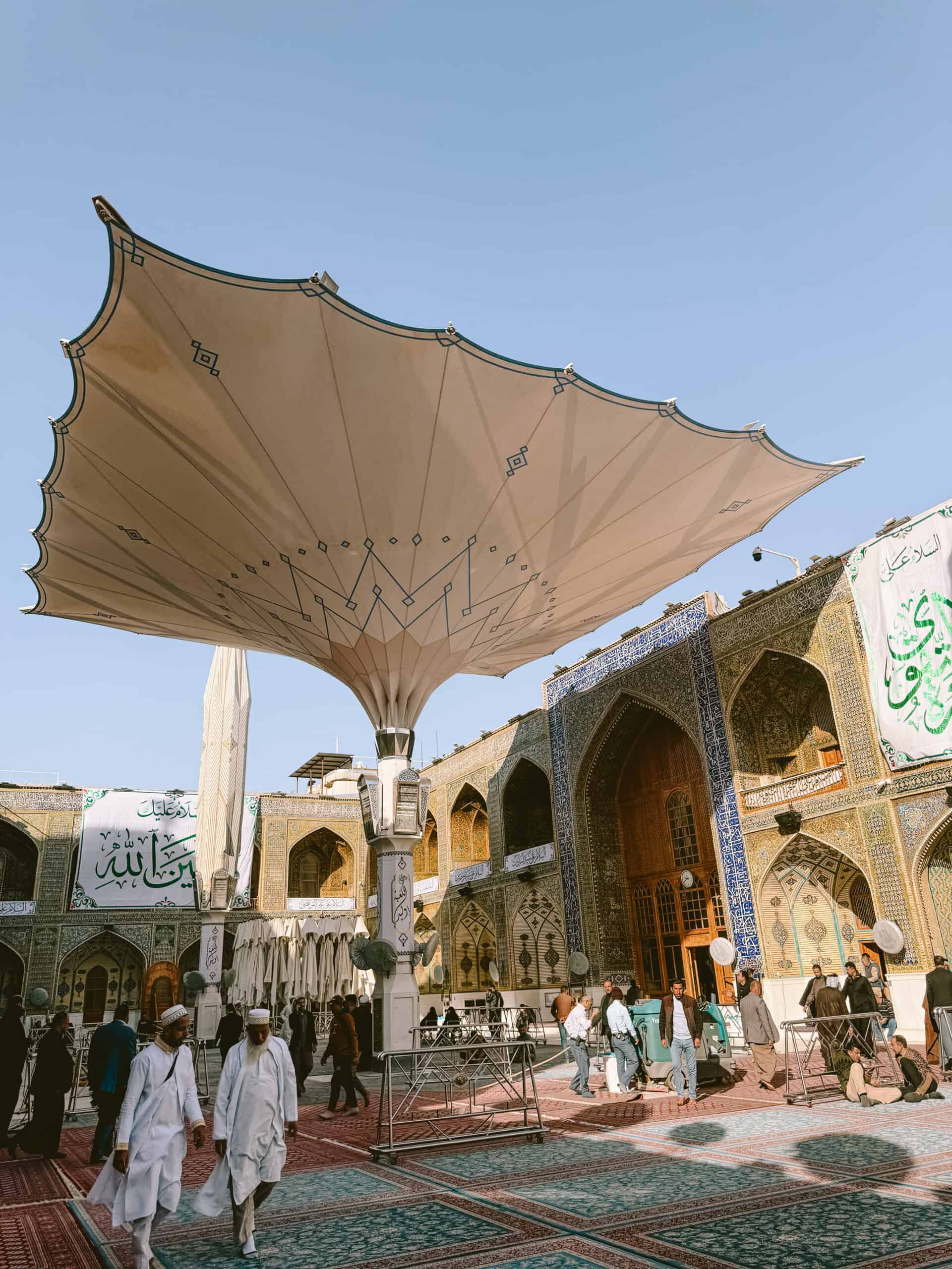 People walk through the colorful tiled courtyard of a shrine, shaded by a massive retractable umbrella structure resembling those found in Medina.