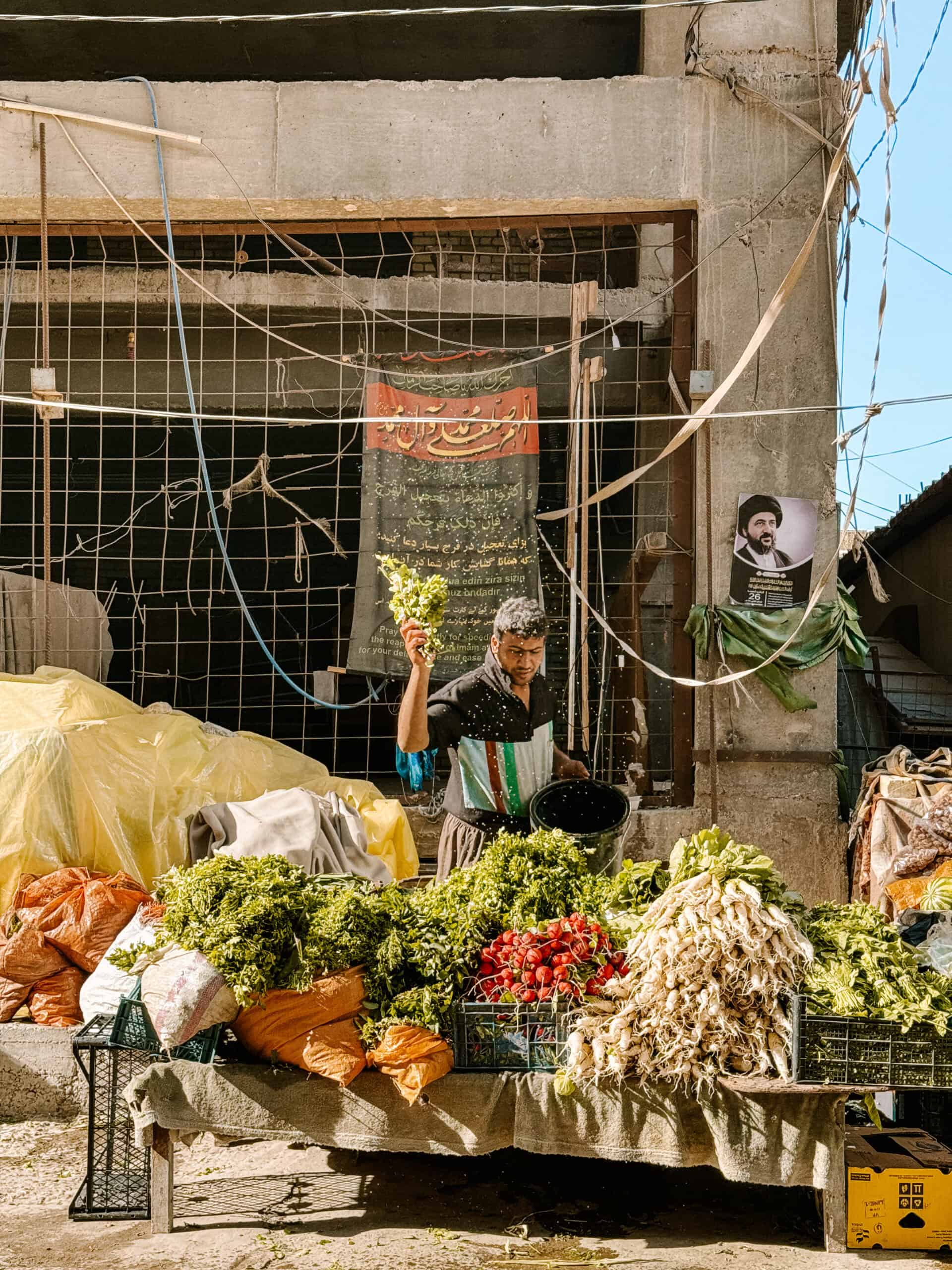 A man in a striped shirt stands behind a table piled with fresh greens, radishes, and herbs, splashing water to keep produce fresh in a local market stall.