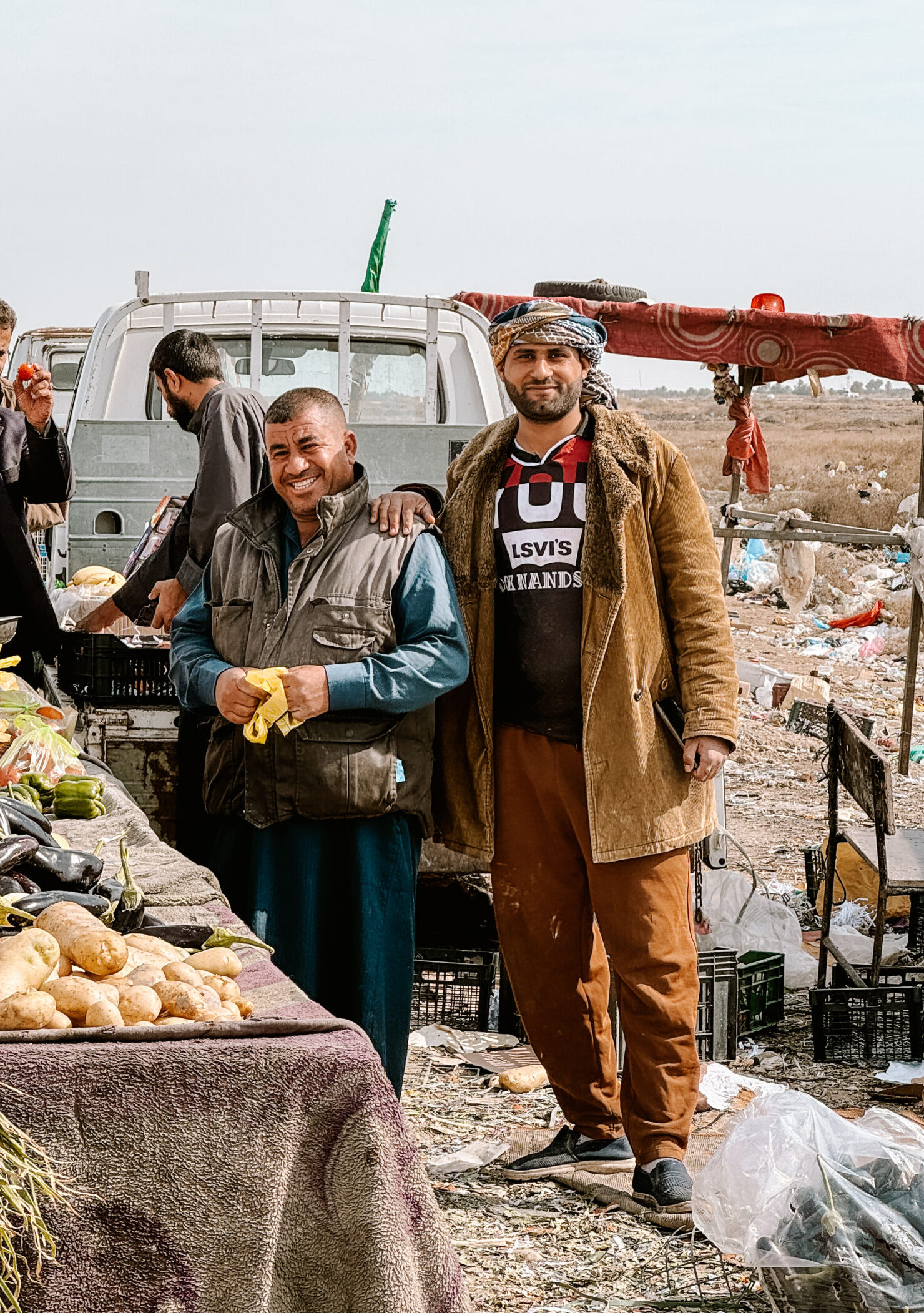 Two men smile beside a rustic produce table filled with potatoes, eggplants, and peppers, in a dusty open-air market surrounded by scattered crates.