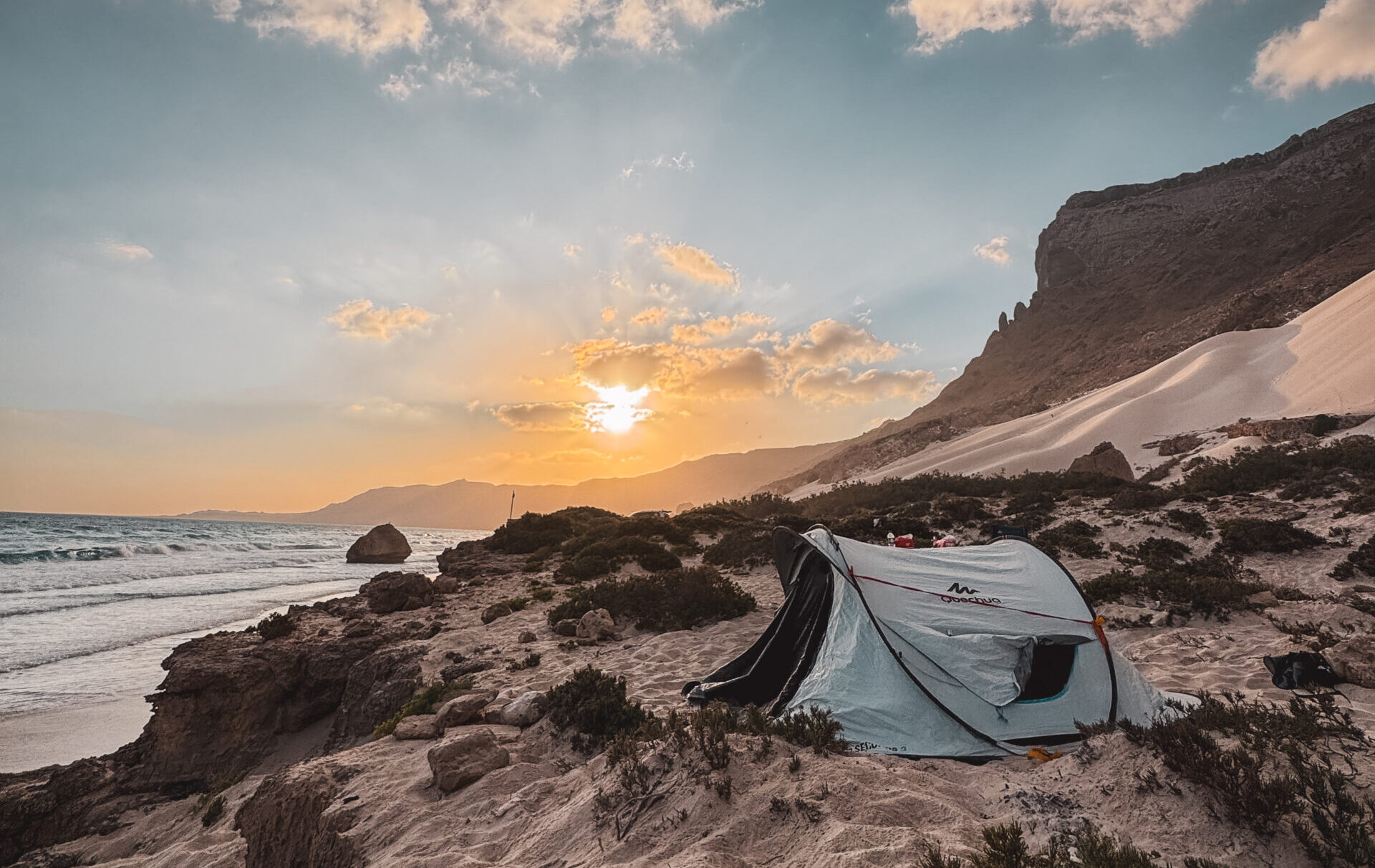 Tent set up on a sandy beach at sunset in Socotra, Yemen, with waves and dramatic cliffs in the background.