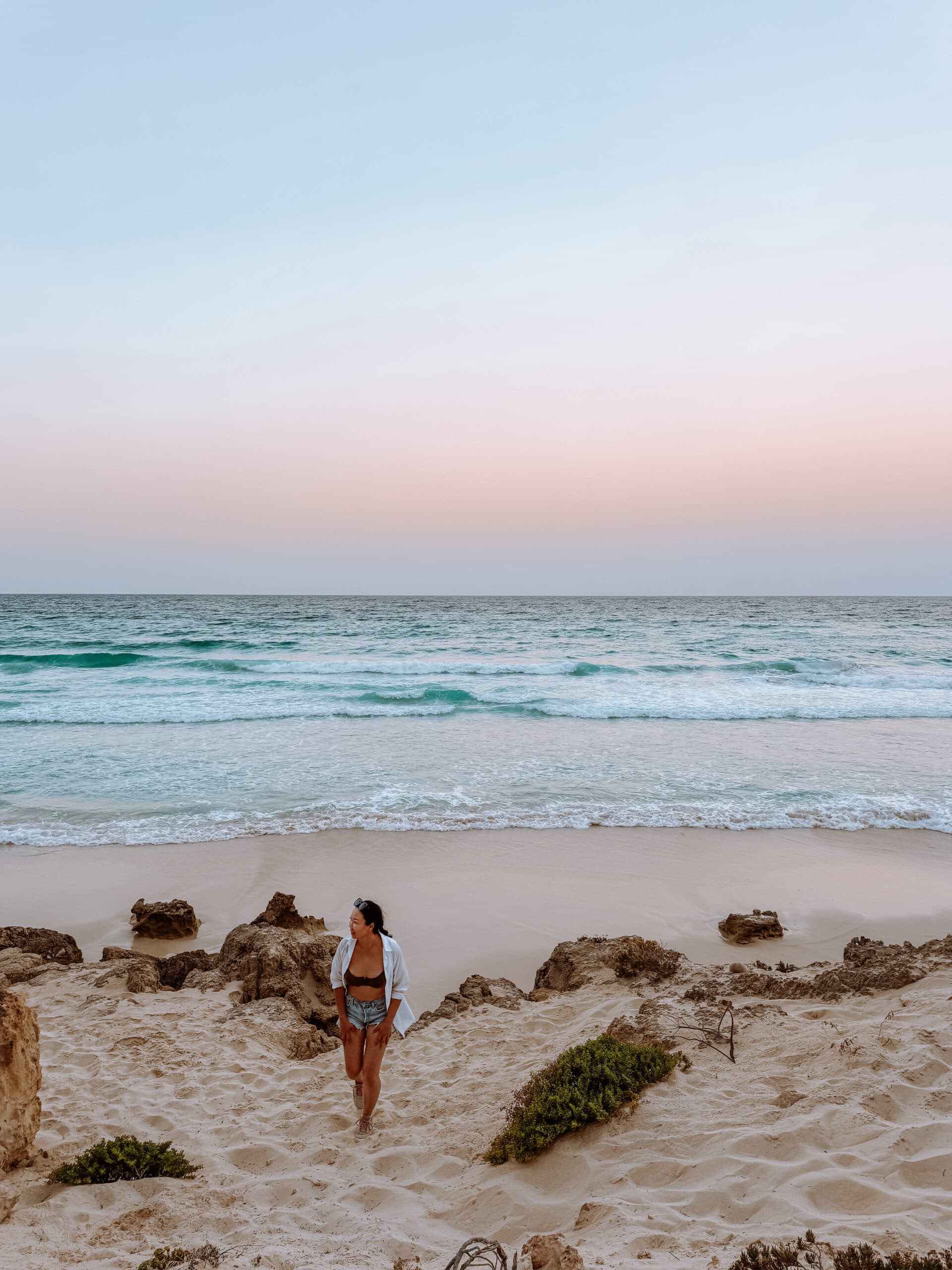 Woman walking on sandy shore at Arher Beach in Socotra during sunset, with soft pink and blue sky over turquoise waves – a serene stop for nature lovers exploring things to do in Socotra.