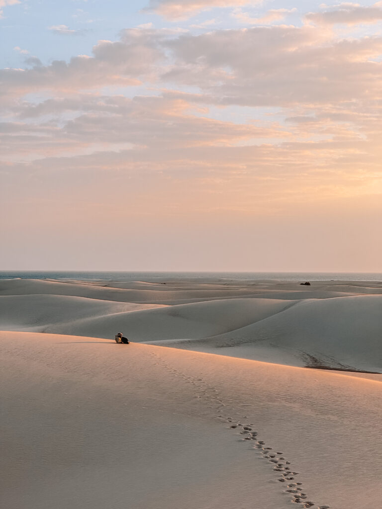 Golden sunrise over the rolling sand dunes of Socotra, with footprints leading to a lone traveler sitting on the crest, a must-see adventure for nature lovers exploring things to do in Socotra.
