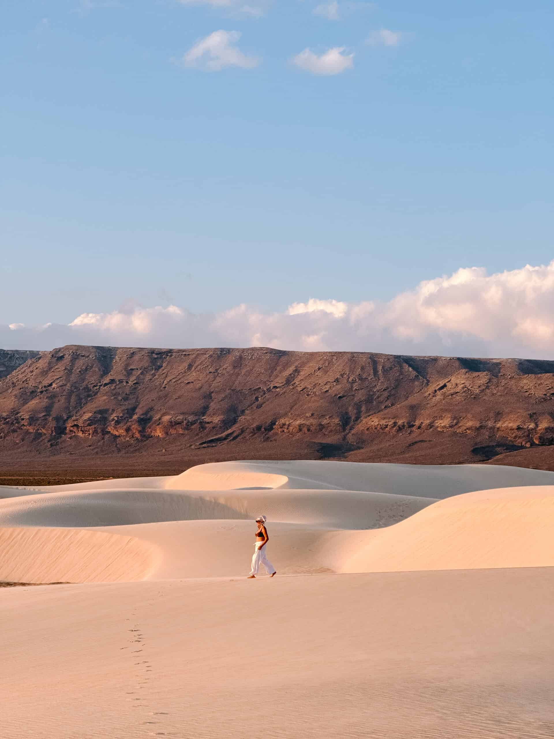 Traveler walking barefoot on Socotra’s white sand dunes at golden hour, with rugged mountains in the background, highlighting the island’s natural beauty and peaceful landscapes.