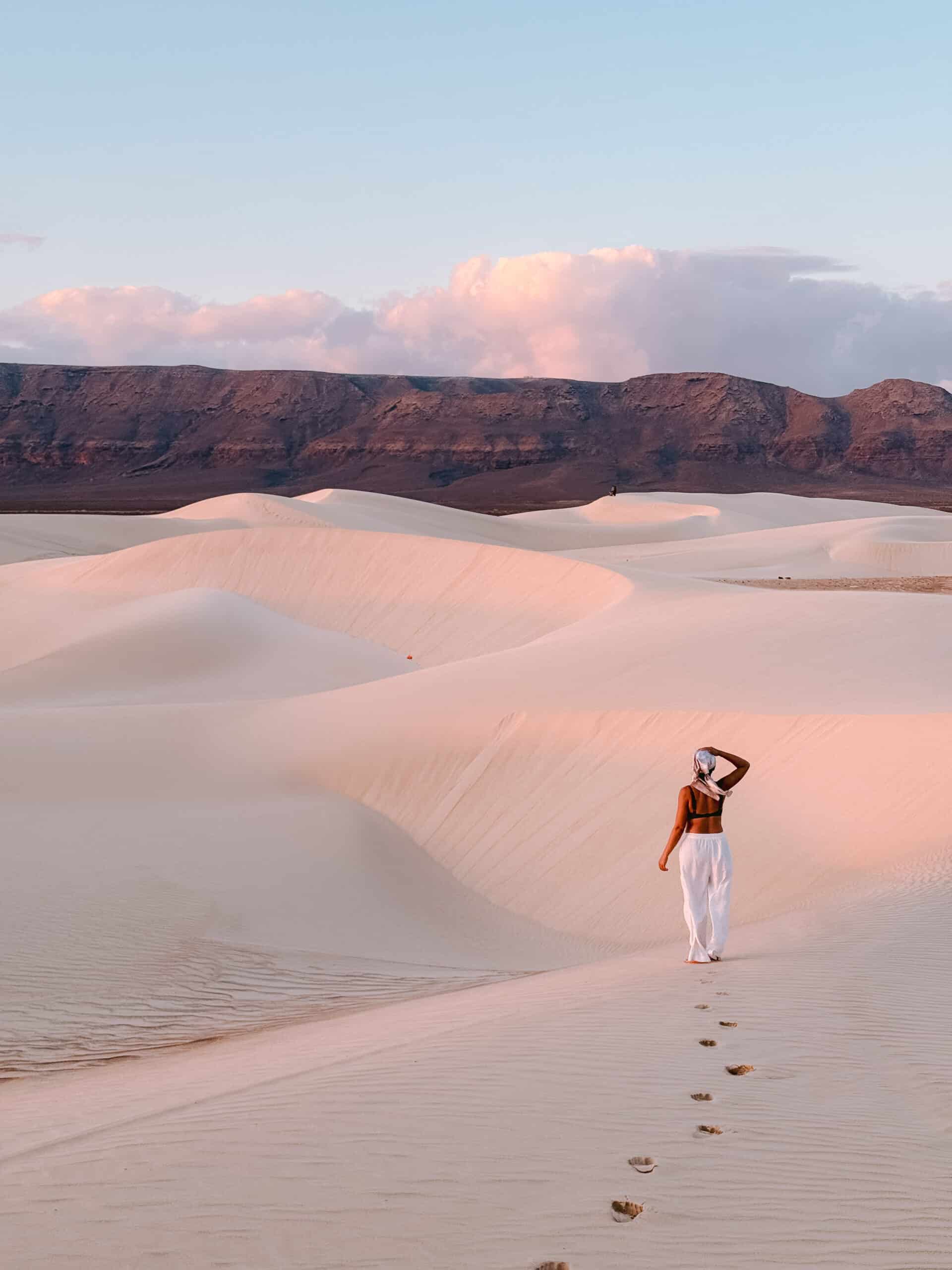 Traveler walking across the golden sand dunes of Zahek in Socotra, Yemen, at sunset with mountains in the background.